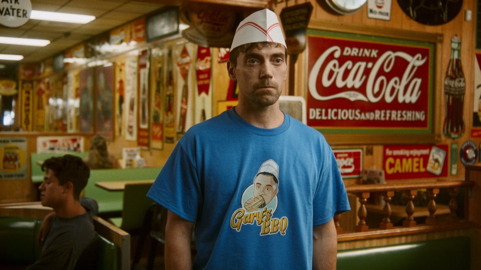 A man wearing a blue t-shirt with 'Gary's BBQ' logo and a paper baseball cap stands inside a vintage-style restaurant decorated with Coca-Cola and other retro signs. A young man is seated at a table in the background.