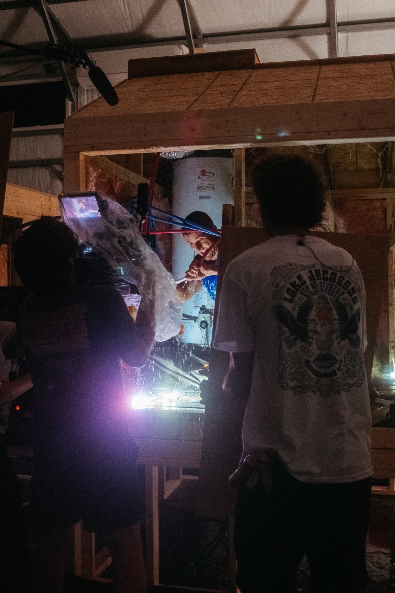 People working on a welding project inside a workshop with wooden framing and a man welding, creating sparks and bright light.