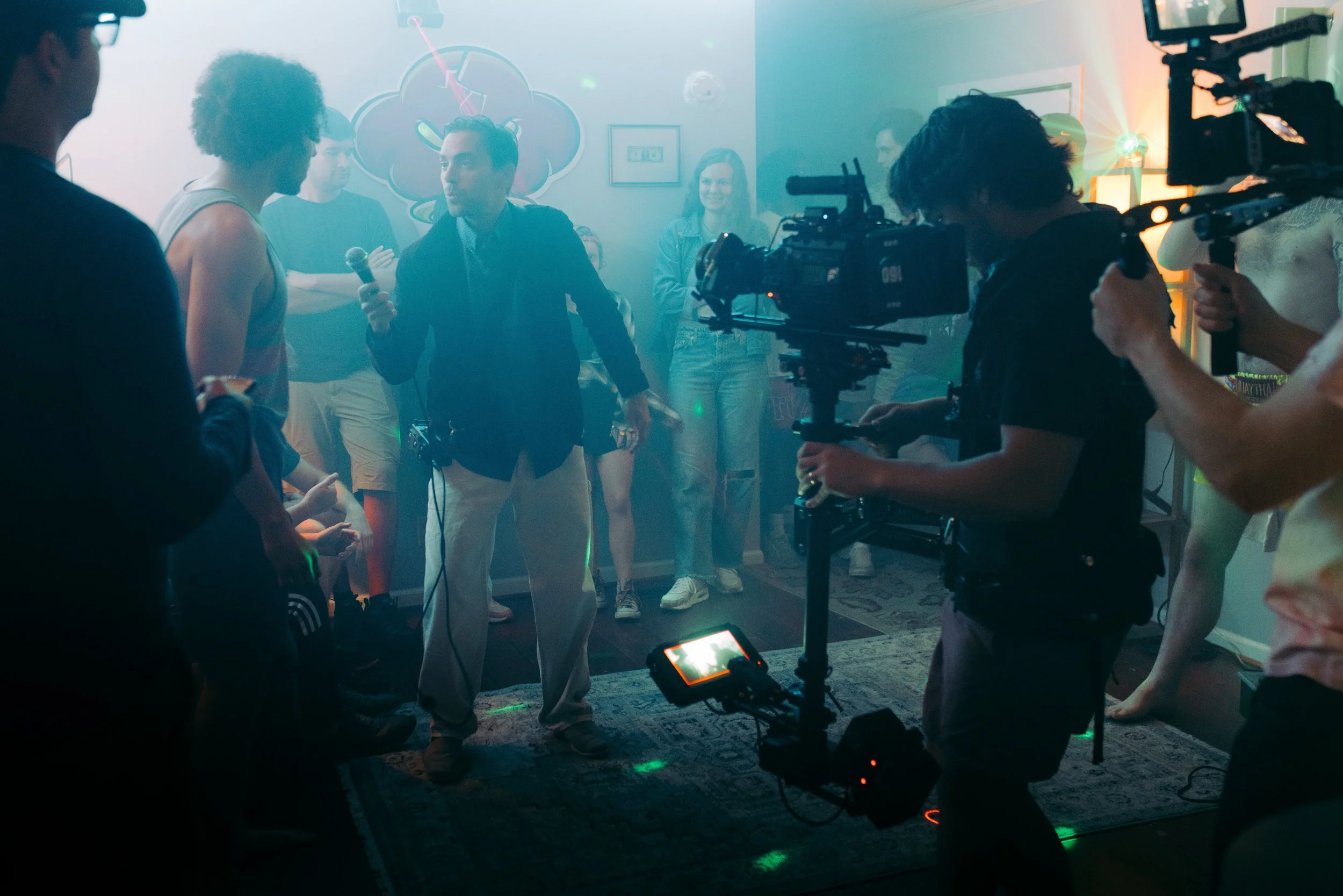 Film crew filming a scene in a party setting with colored lights and a fog machine, a woman holding a microphone, and several people watching.