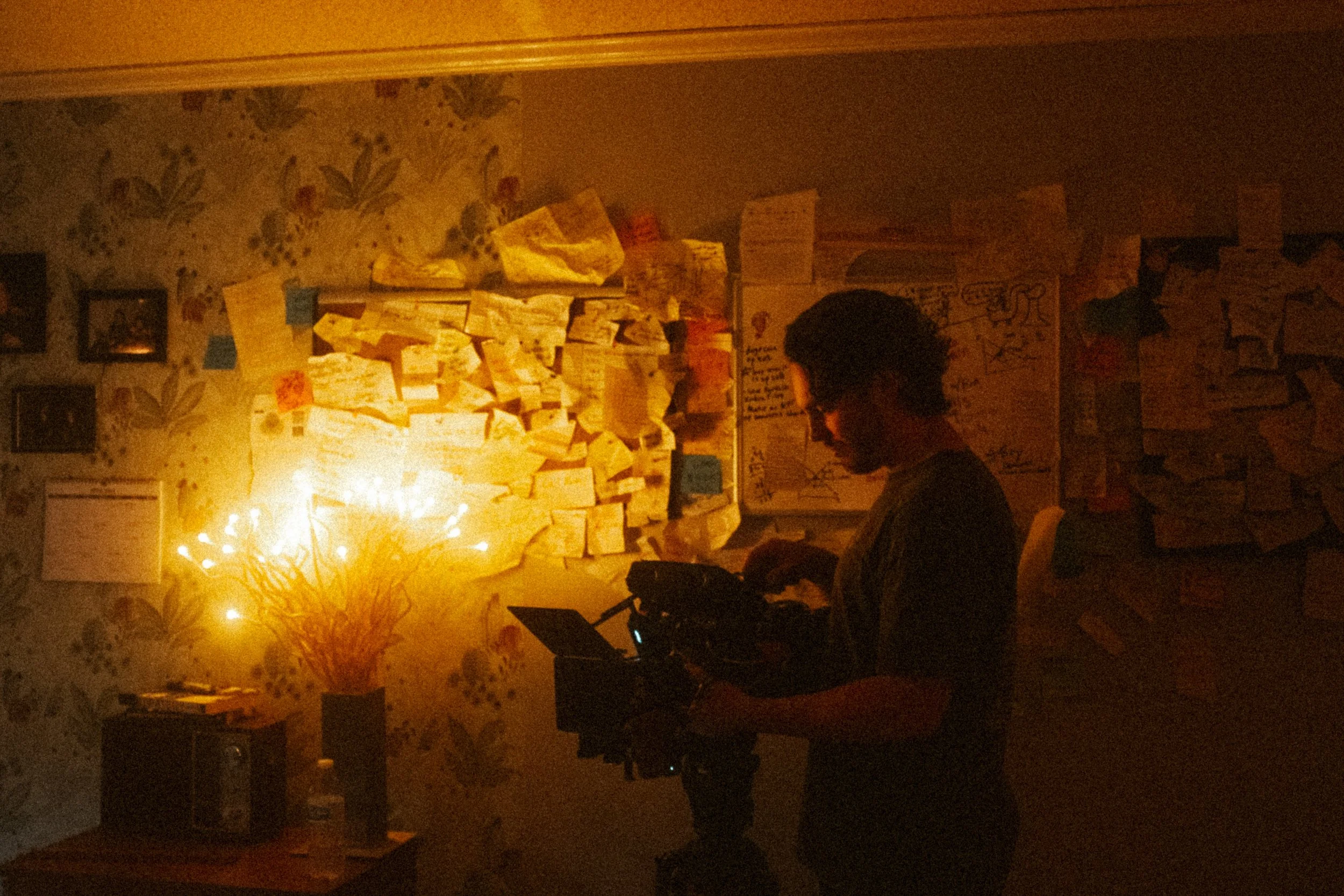A man operating a camera in a dimly lit room with wallpaper, notes, and papers on the walls, and a small table with a water bottle and a speaker.