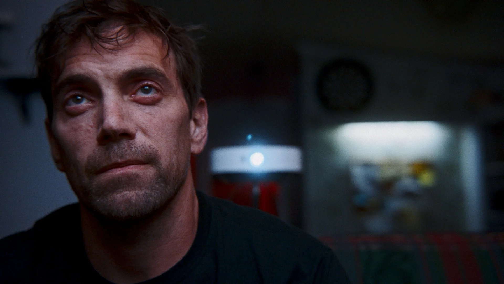 Close-up of a man with short, brown hair and stubble, looking upward with a neutral expression. The background shows a blurry object resembling a clock and a light source.