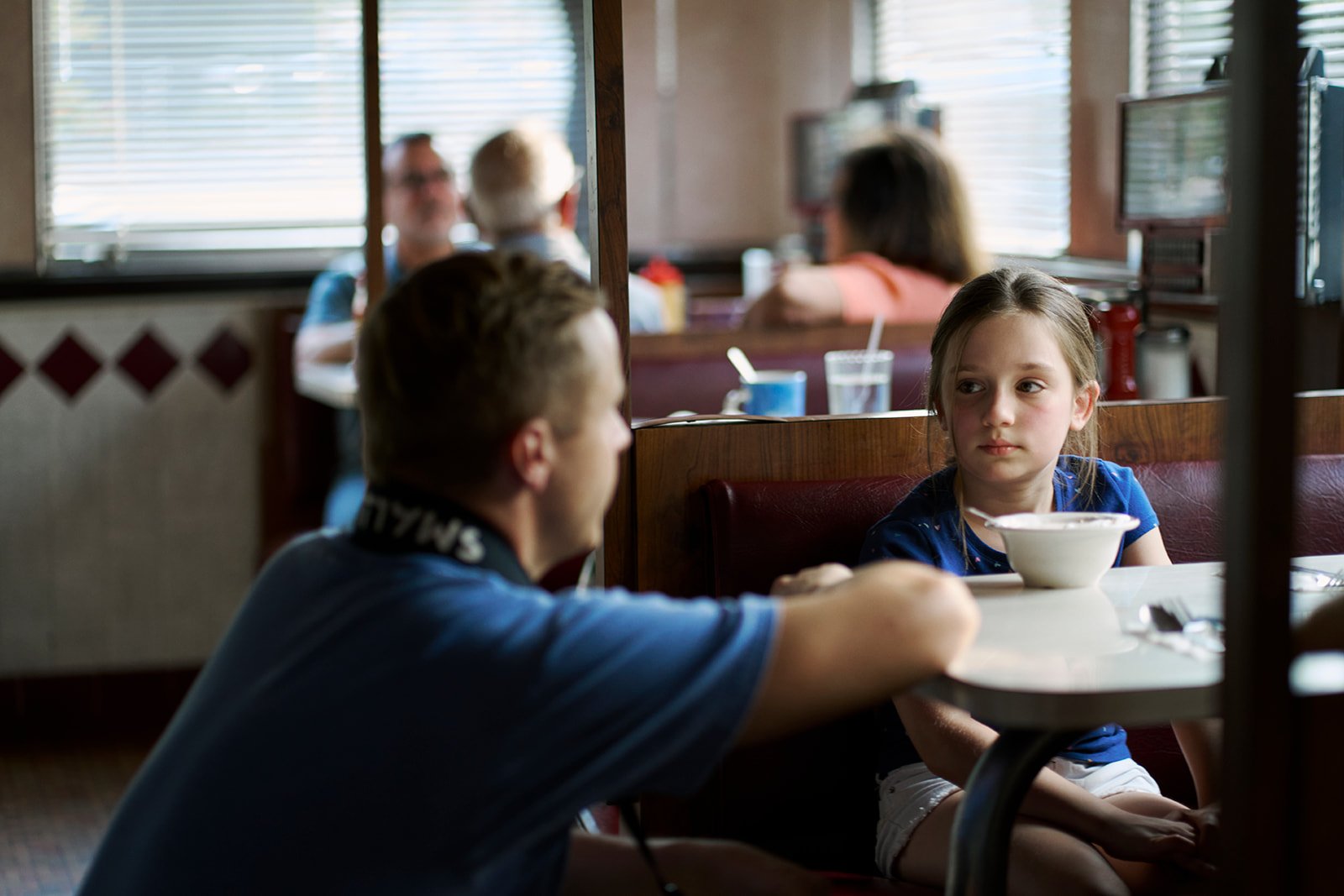 A young girl and a man are sitting at a booth in a restaurant. The girl is looking at the man with a serious expression, and there is a bowl of food in front of her. The restaurant has wooden booths and large windows letting in natural light, with other customers in the background.
