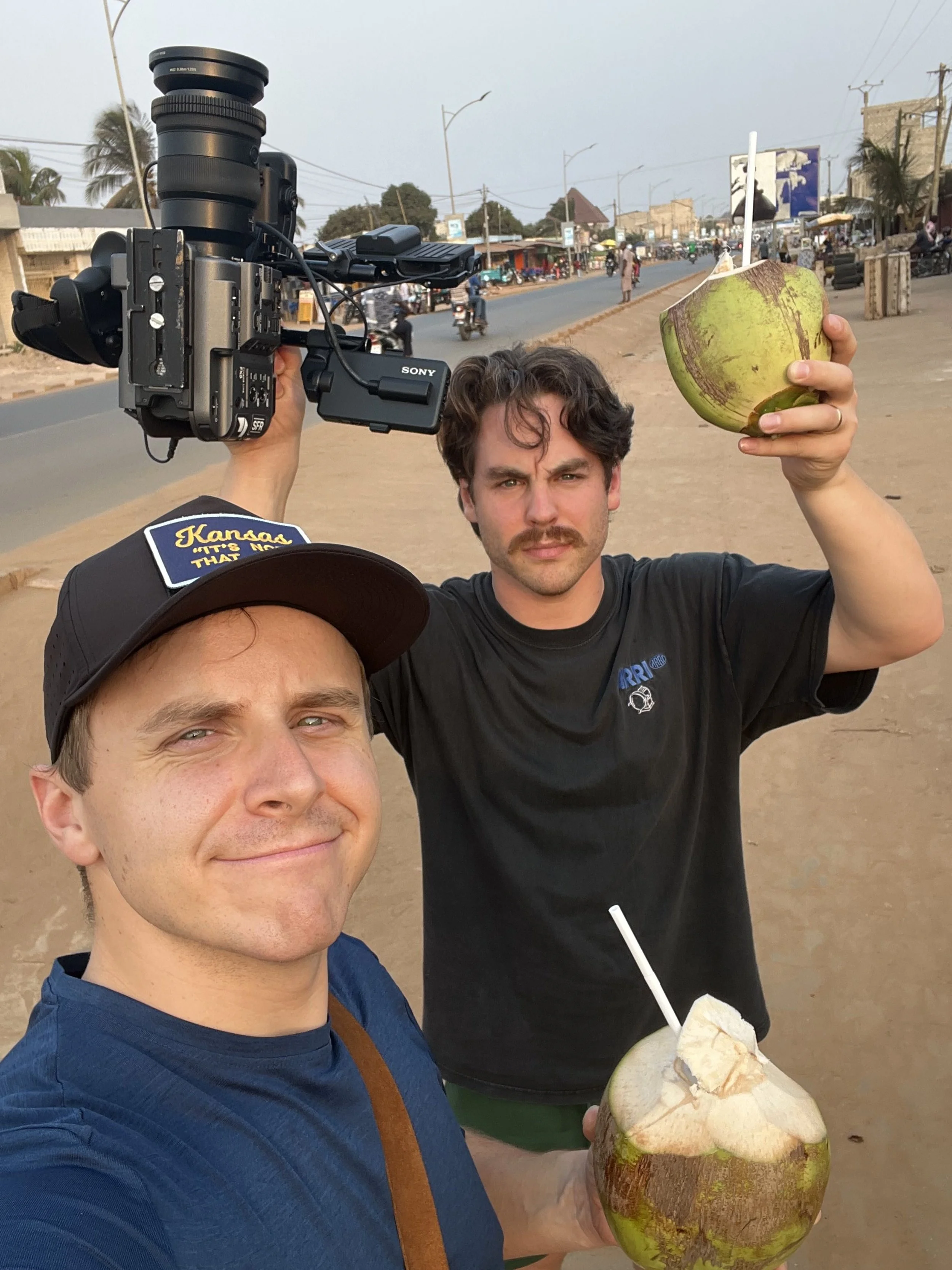 Two men taking a selfie on a street, each holding a green coconut with a straw. One man is smiling at the camera, while the other is holding a video camera and has a serious expression.
