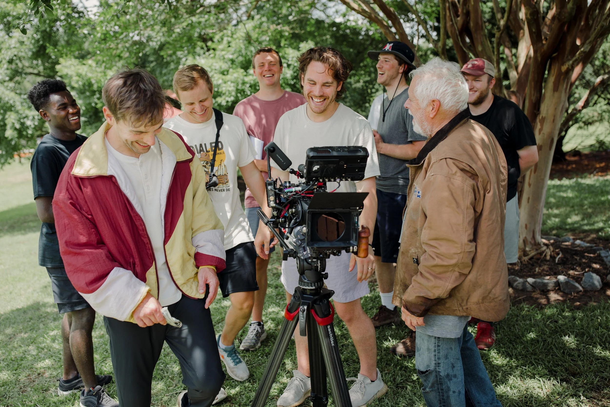 Group of people on a film set outdoors around a camera, smiling and laughing, with trees in the background.