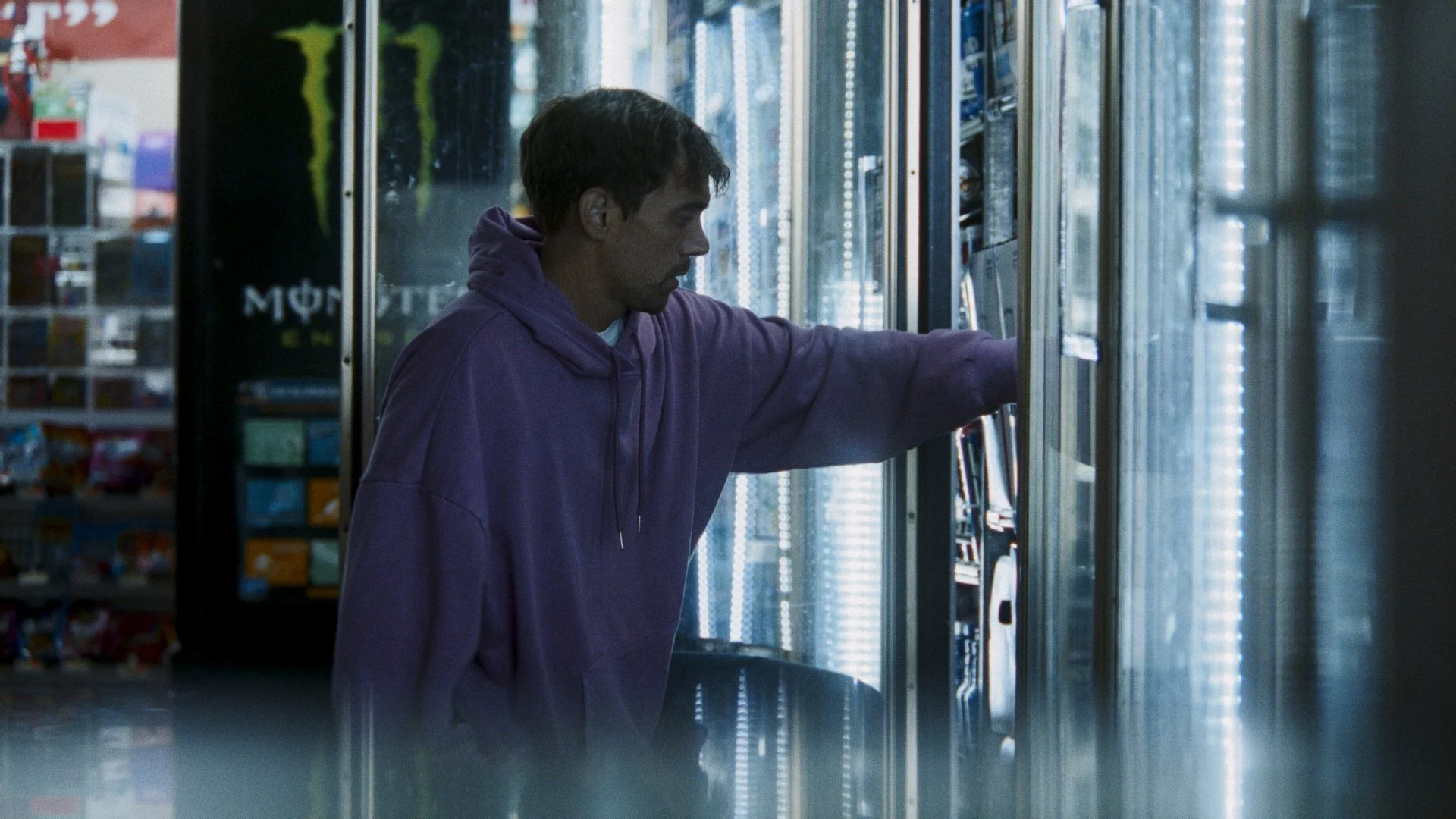 A man in a purple hoodie reaching into a vending machine in a store, with snack shelves visible in the background.