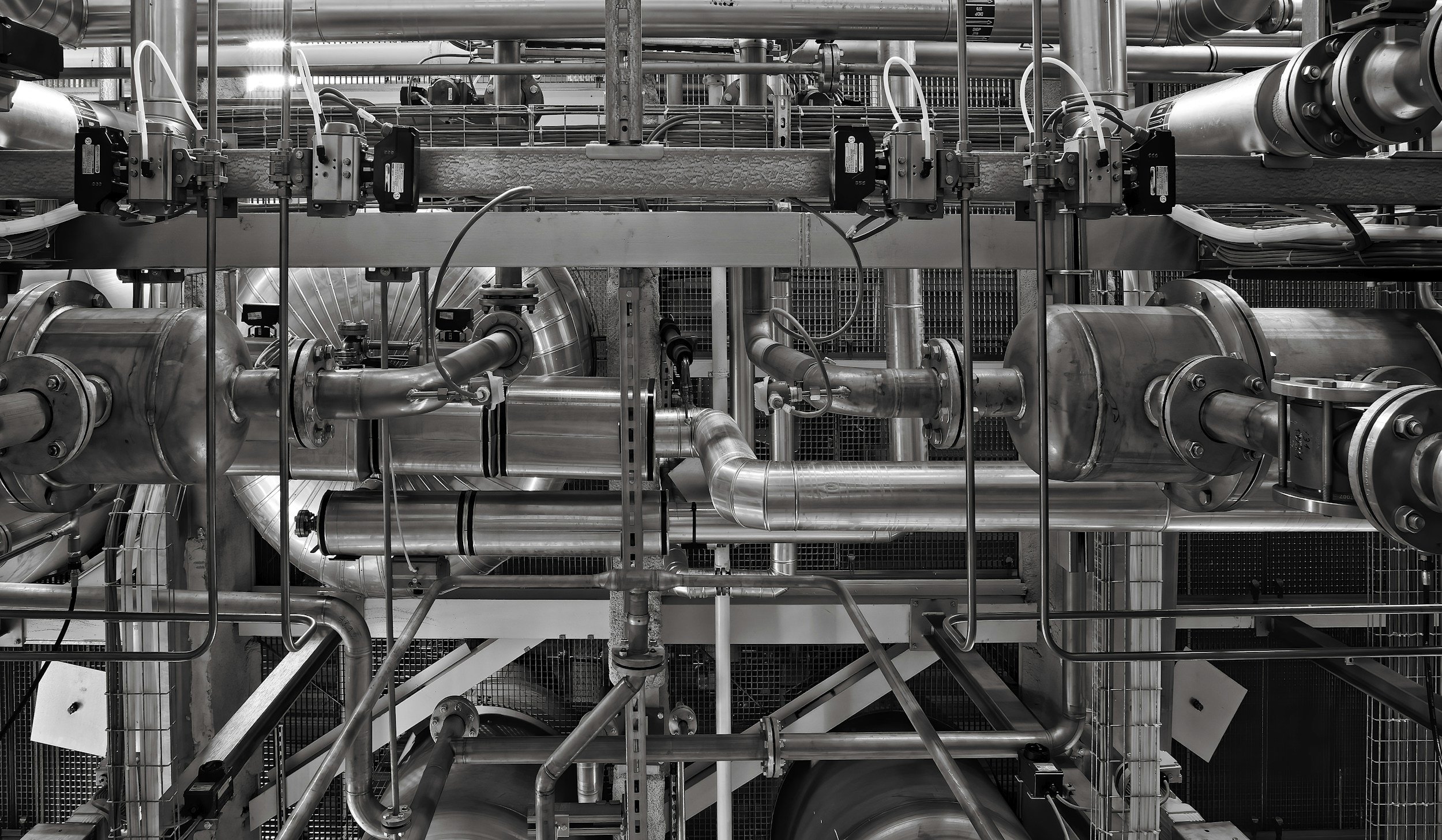 A complex network of stainless steel pipes, valves, and machinery in an industrial setting, possibly part of a factory or processing plant.