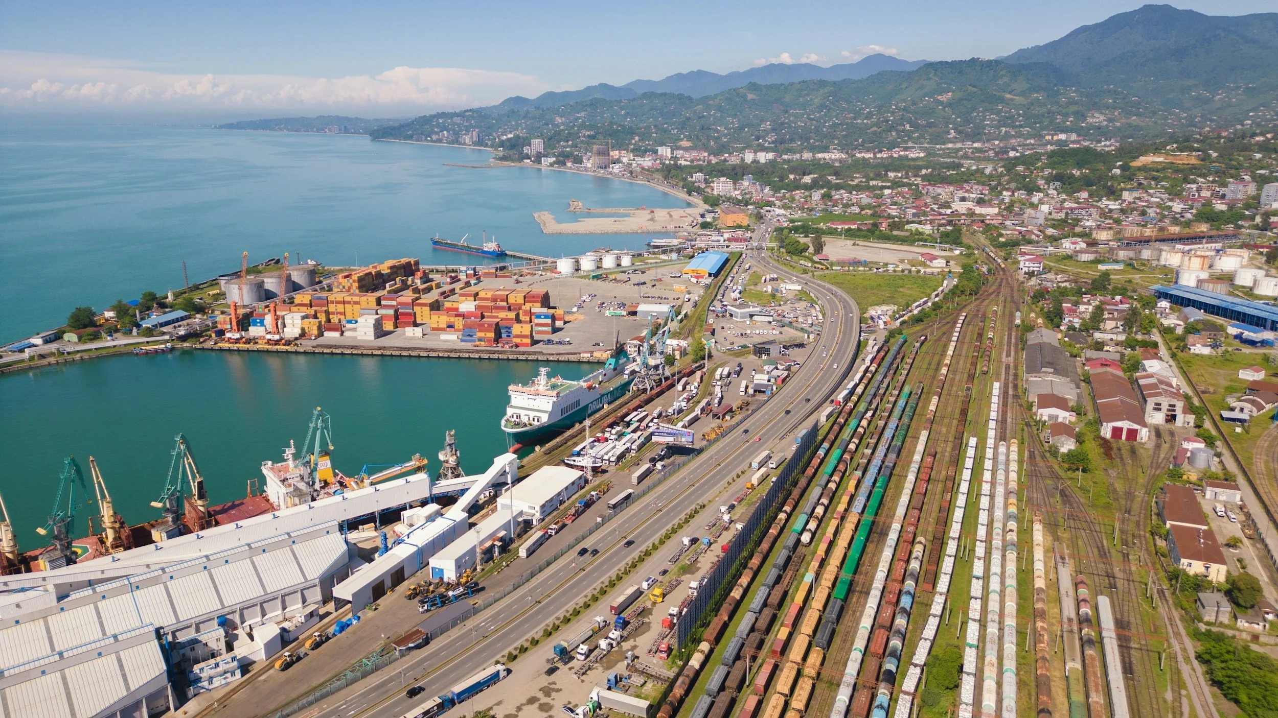 Aerial view of a busy port with cargo ships, containers, trucks, train tracks, and a city with mountains in the background along the coastline.