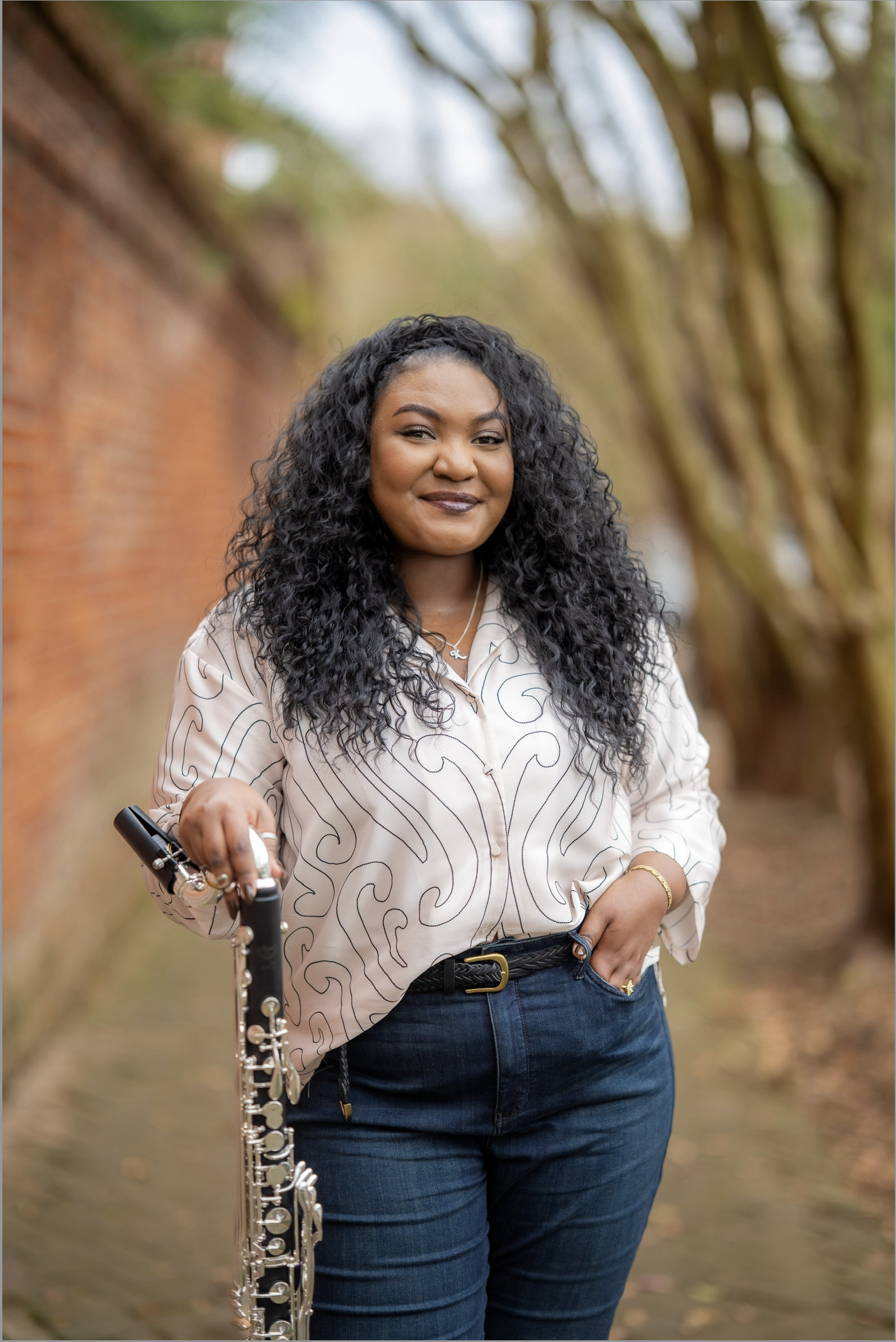 A woman with curly black hair smiling at the camera, holding a clarinet in her right hand, standing outdoors on a path lined with trees and a brick wall.