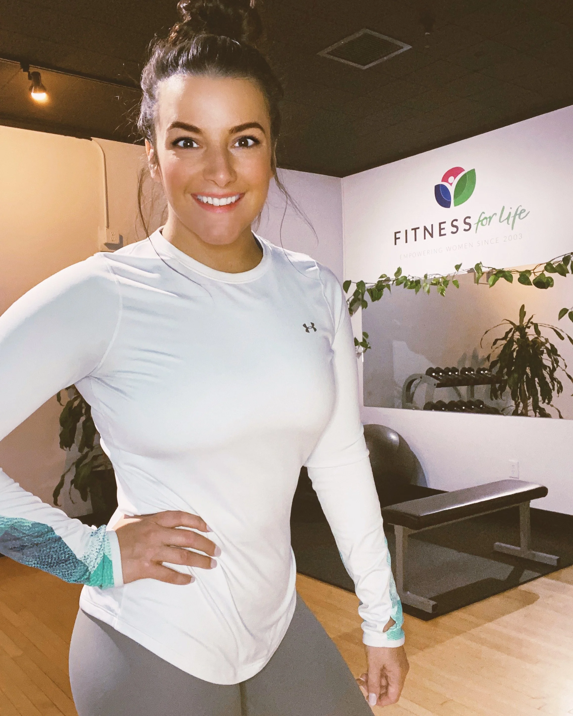 A smiling woman in workout attire standing inside a fitness studio with a sign that reads 'Fitness for Life' on the wall behind her.