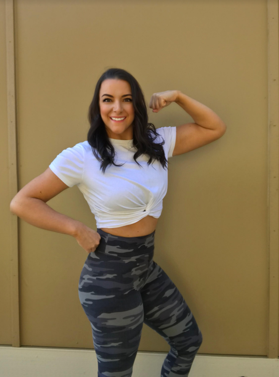 A woman with dark hair smiling, wearing a white knotted t-shirt and camouflage leggings, flexing her right arm and standing in front of a plain beige wall.