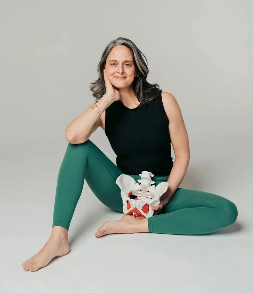 A woman with gray hair sitting in a cross-legged position, holding a pelvis model, wearing a black sleeveless top and green leggings, smiling softly against a plain light background.