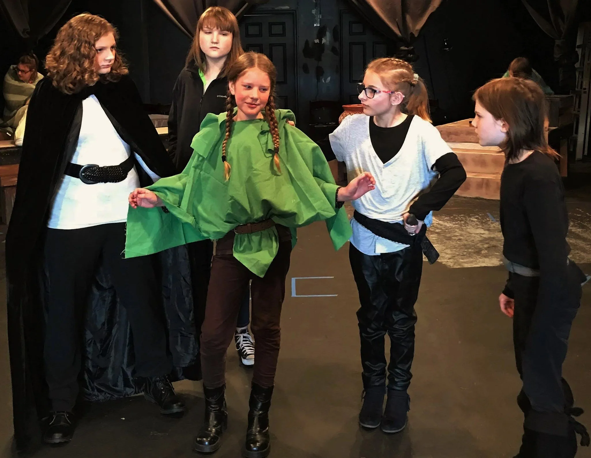 A group of children in costume, with two girls in the center wearing green and black outfits, interacting on a stage or theater setting.