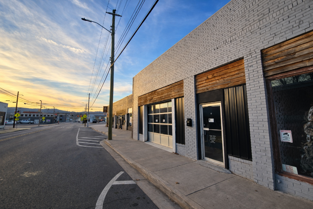 Empty street with sidewalk and storefronts under a sunset sky, with utility poles and power lines along the road.