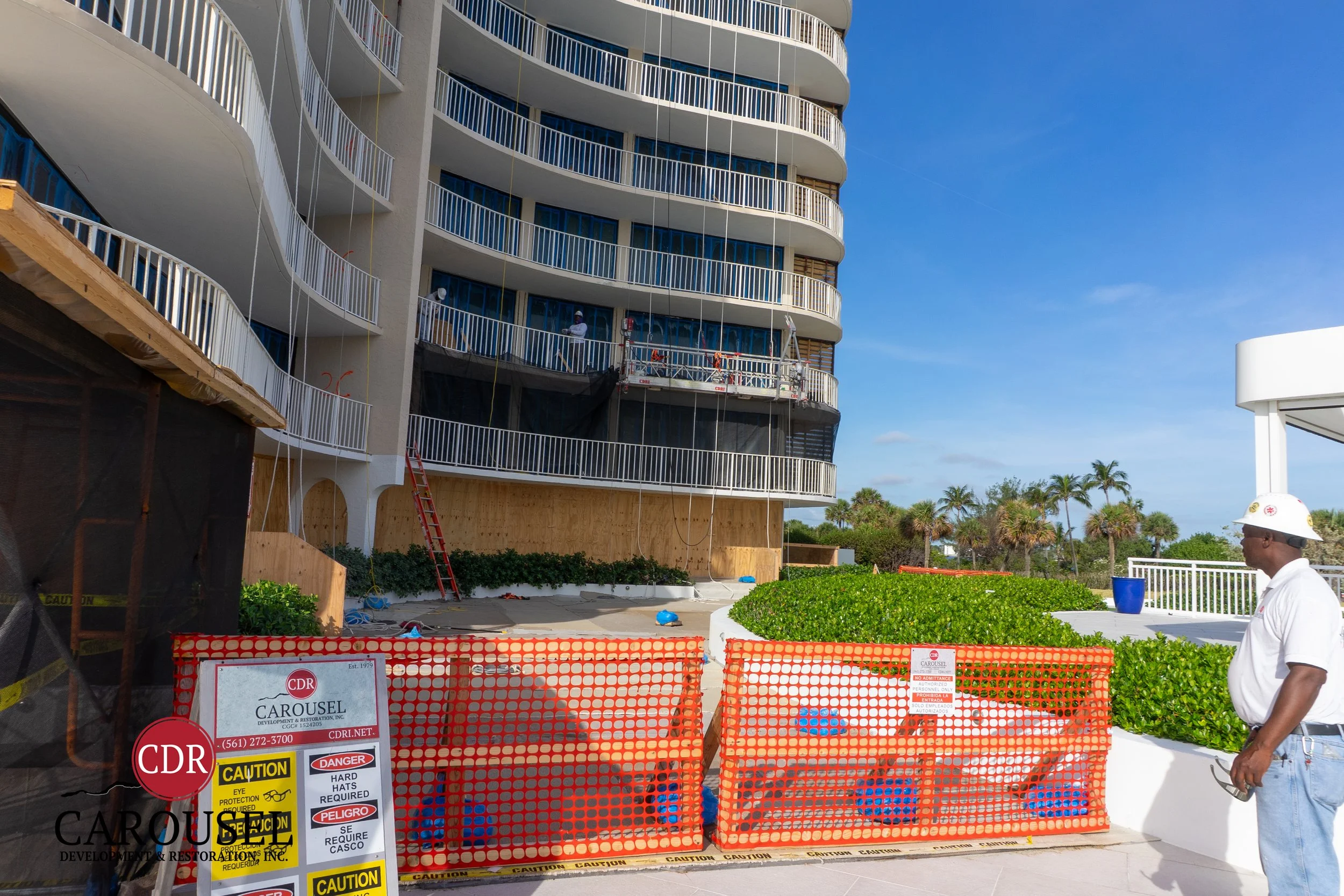 Construction workers on scaffolding in front of a multi-story building with curved balconies, outside beach area with palm trees, safety orange fencing and caution signs.