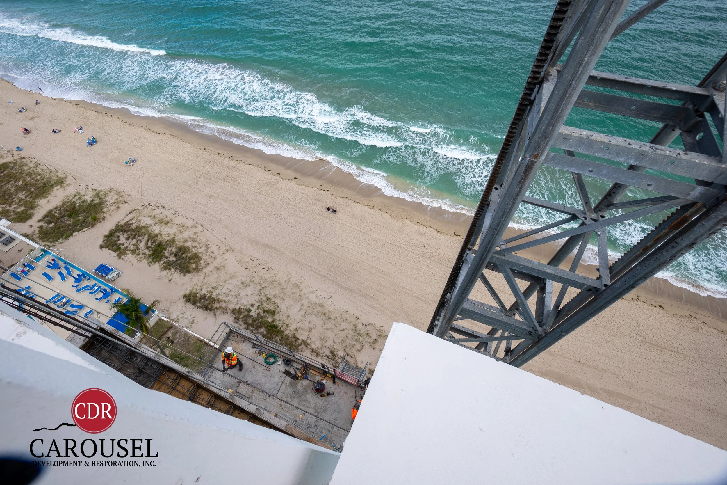 Construction workers working on a building by the beach, with the ocean and waves visible in the background from a high vantage point.