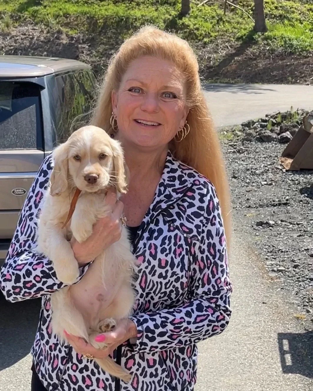 A woman with long blonde hair, wearing a leopard print jacket, holding a cute tan puppy with floppy ears outdoors next to a gray vehicle.