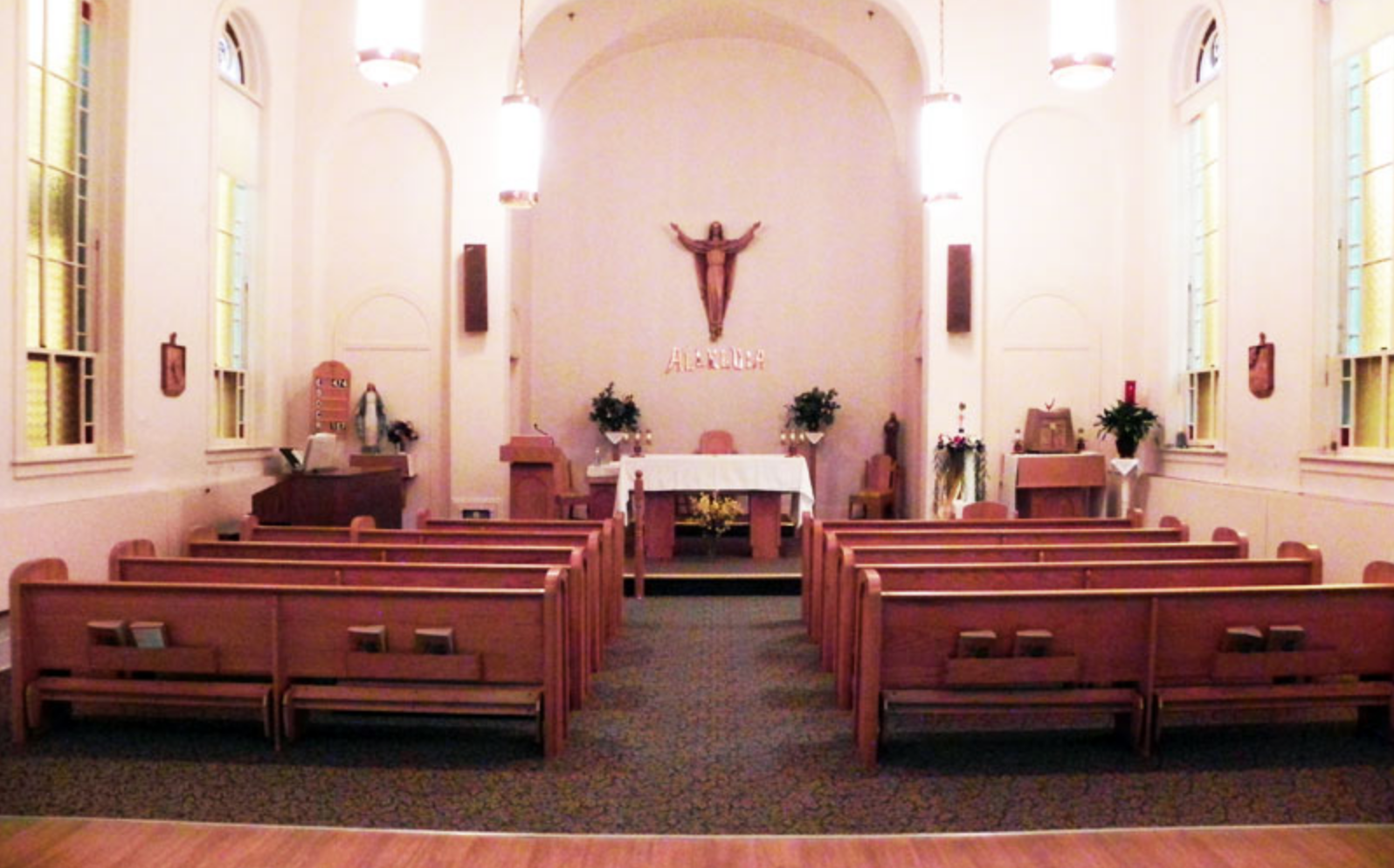 The interior of a small church with wooden pews, a white altar, and religious decorations, including a crucifix with Jesus on the wall behind the altar.
