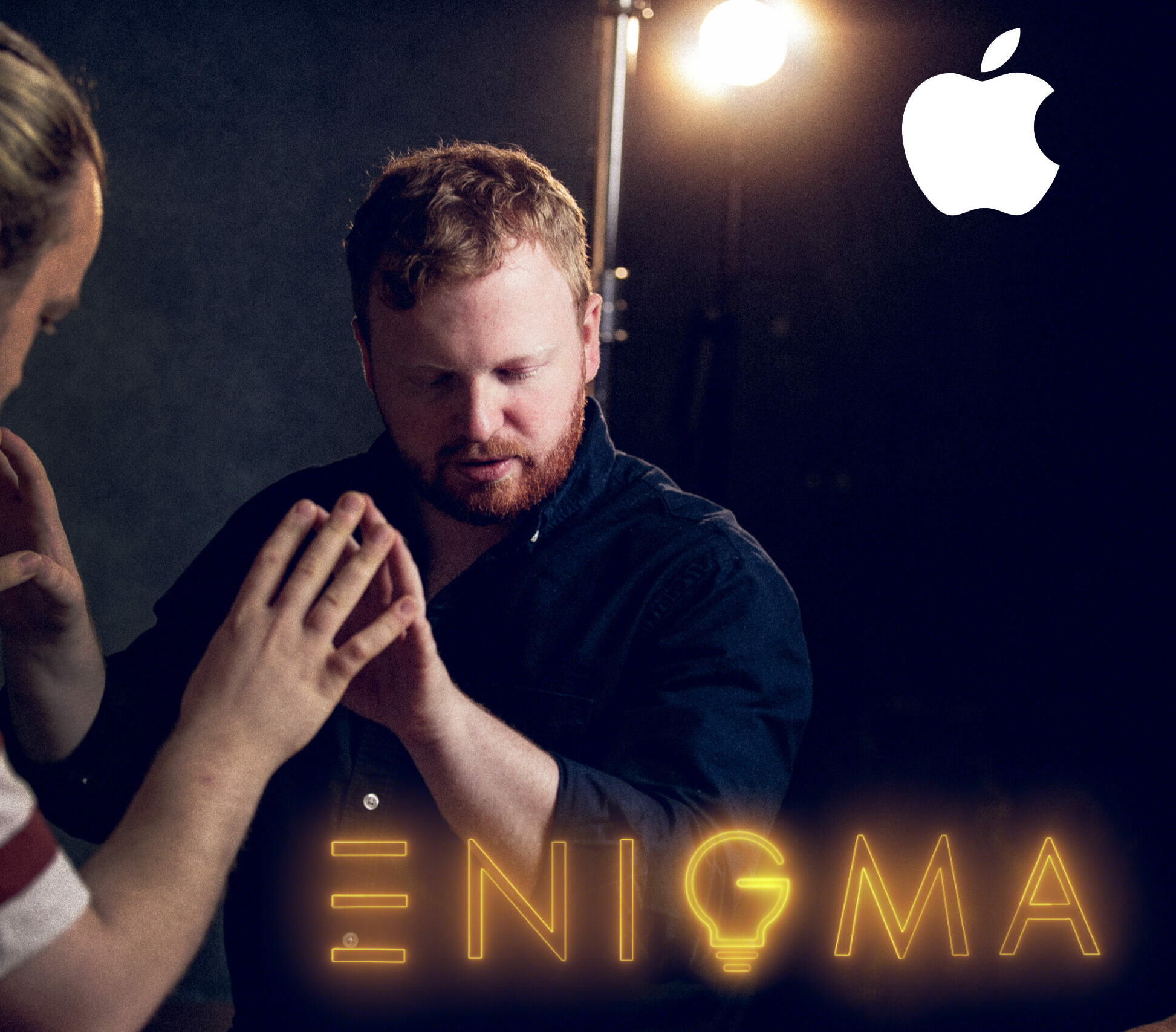 A man and a woman holding hands together, with the woman inside a dimly lit room. The image features a neon sign that reads 'ENIGMA' with a lightbulb icon inside the letter 'G' and an Apple logo in the top right corner.