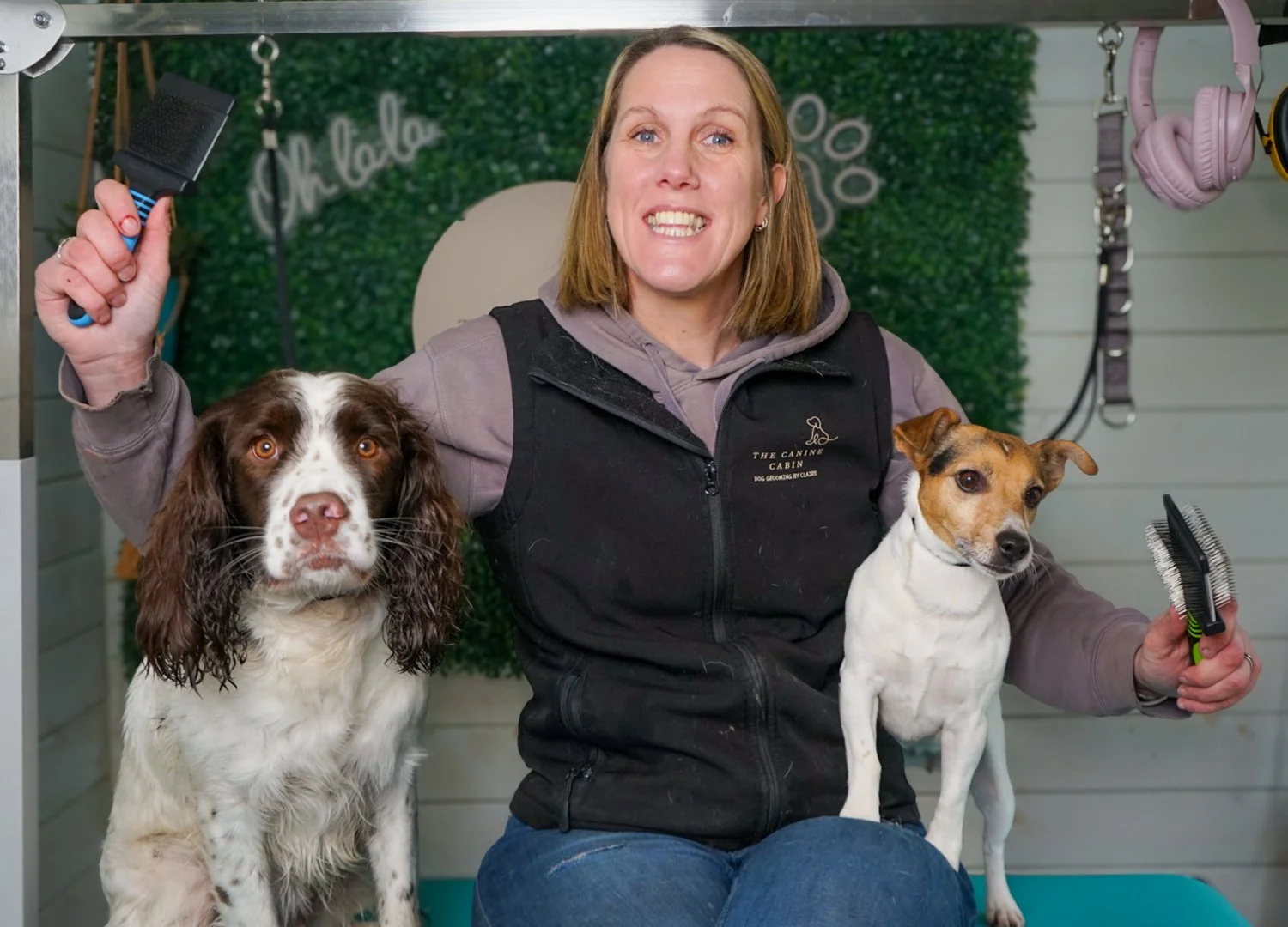 A woman with two dogs sitting on her lap, holding grooming brushes, in a pet grooming salon.