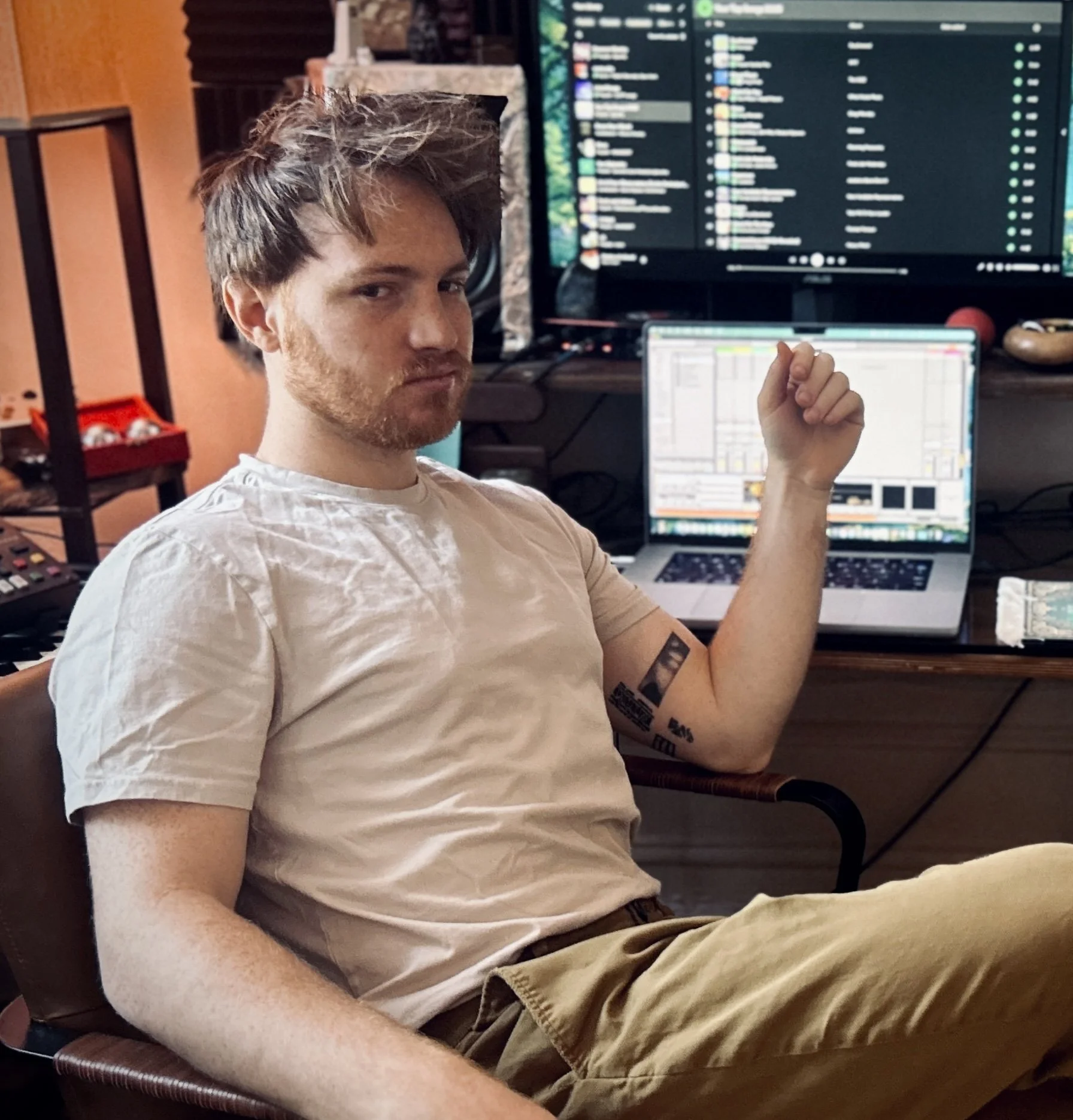 A young man with disheveled hair and a beard, sitting in a chair in front of a desk with computer screens, making a fist pump gesture with his right hand and looking at the camera.