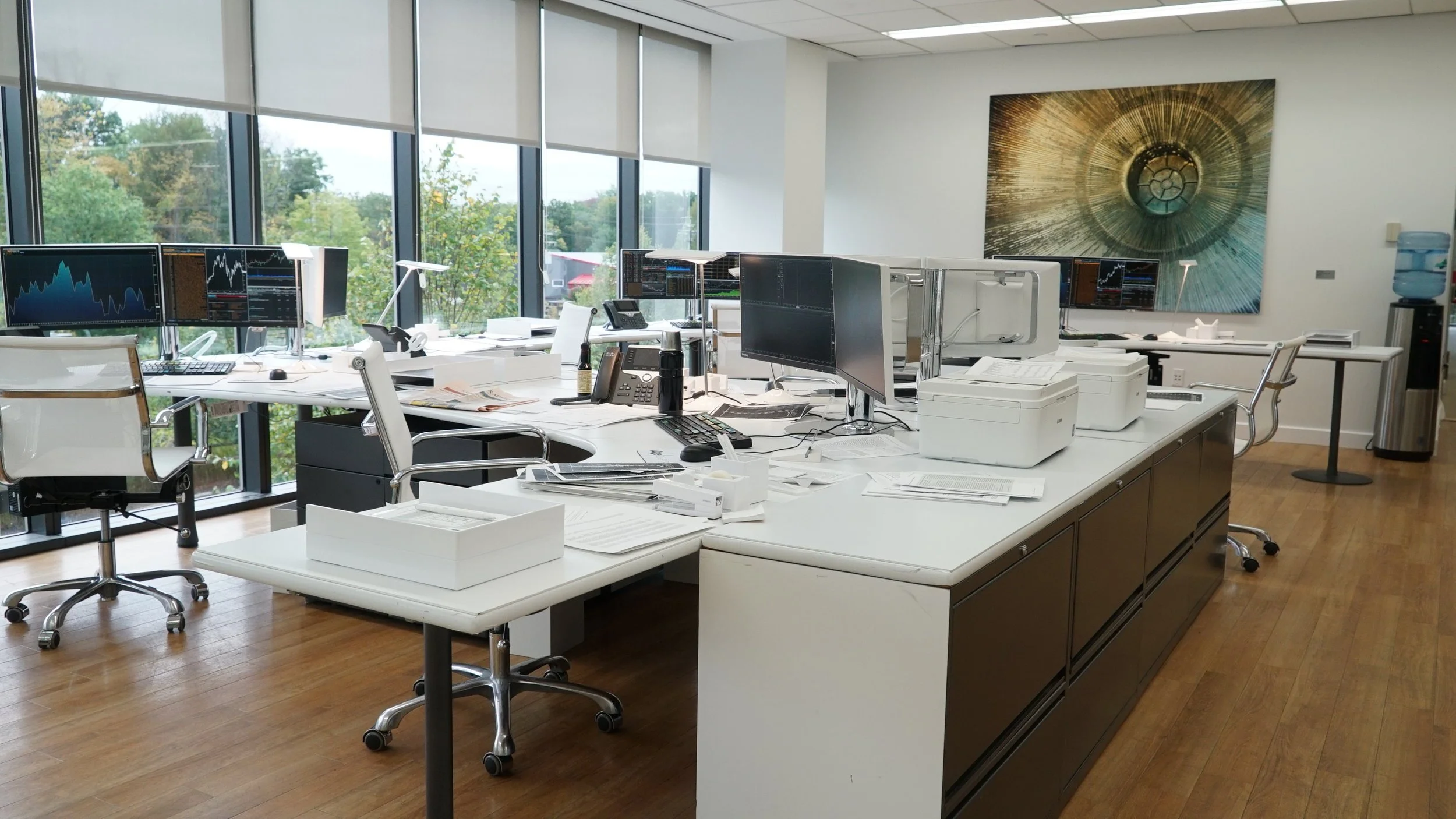 Empty office workspace with multiple computer monitors, desks, chairs, office supplies, and a water cooler. Large windows with blinds reveal greenery outside.