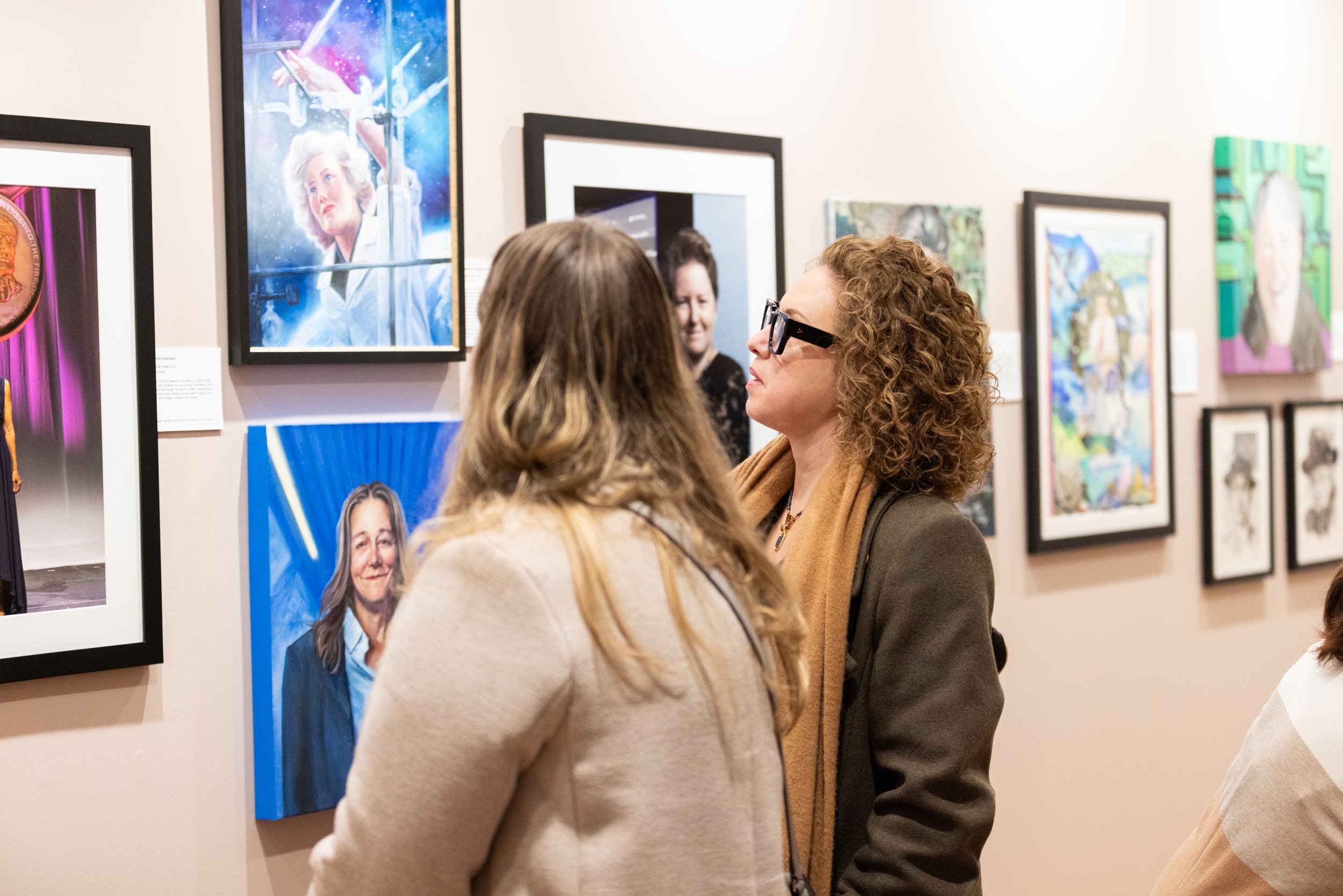 Two visitors exploring paintings and portraits of women and gender expansive people at The Alcove Portrait Gallery, a feminist art space in Providence, RI