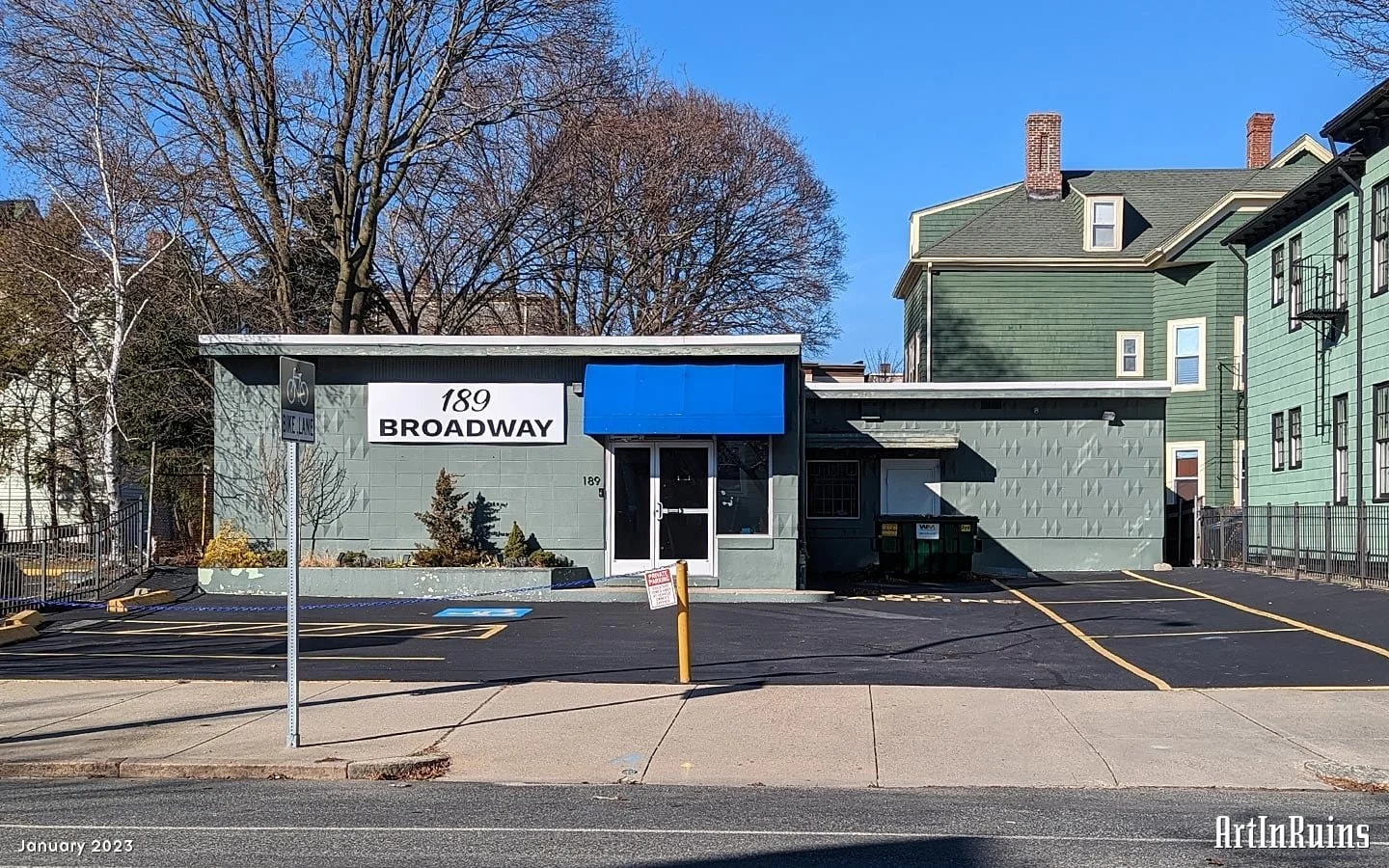 Single-story commercial building at 189 Broadway with light green exterior walls, white trim, and a blue awning over the entrance. The building has a small parking lot and is situated in a residential area with bare winter trees in the background.