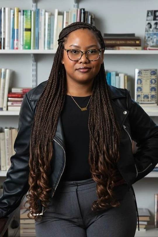 Professional portrait of a woman with long braided hair wearing glasses, a black leather jacket, and dark clothing, standing in front of white bookshelves filled with books in an office or library setting.