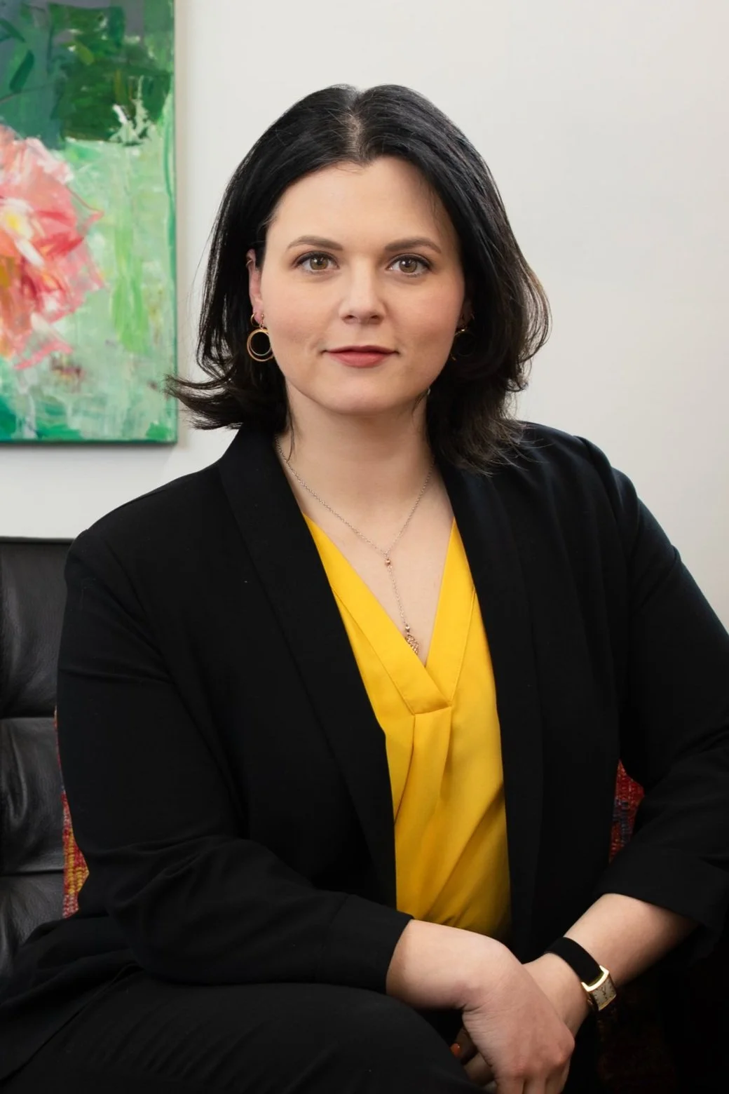 Professional headshot of a woman with shoulder-length dark hair wearing a black blazer over a yellow top and circular earrings, photographed against a white background with a colorful abstract painting visible to the side.