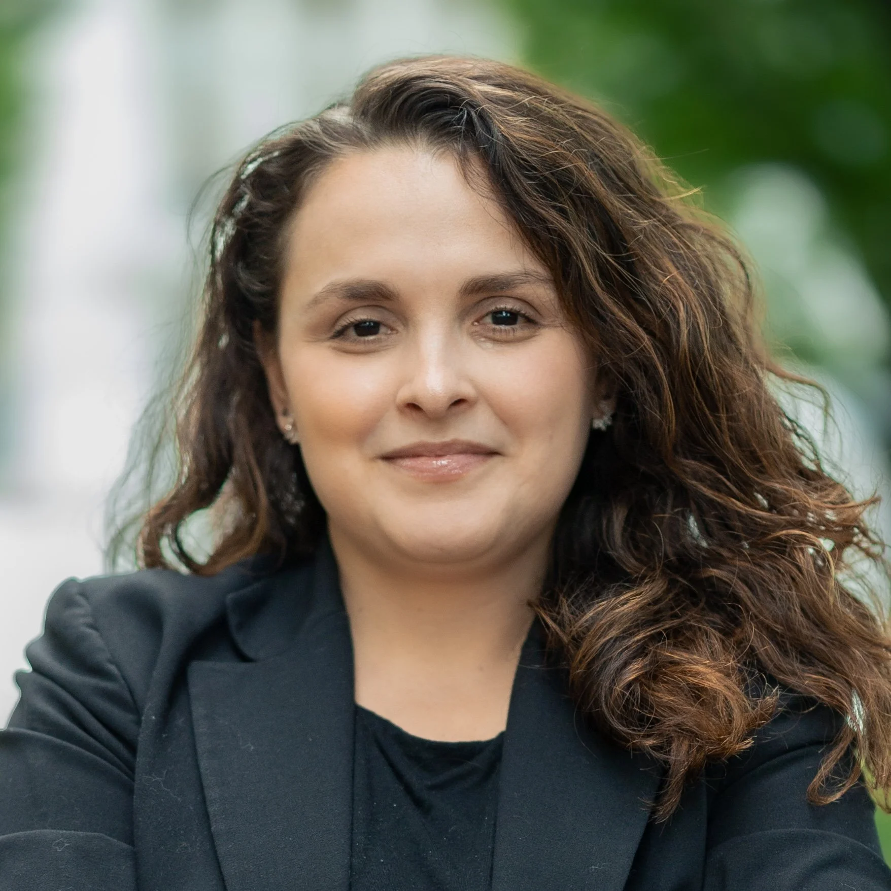 Professional portrait of a woman with long wavy brown hair wearing a dark blazer and small stud earrings, photographed outdoors with a soft blurred green background.
