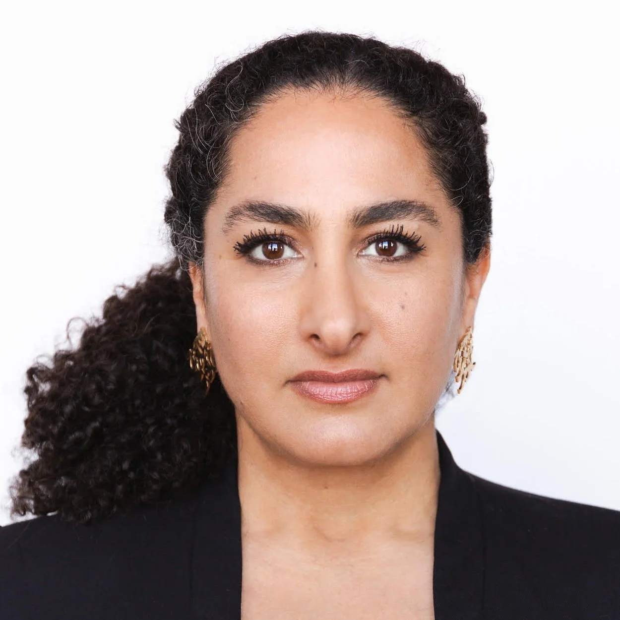 Headshot of a woman with dark curly hair pulled back, wearing gold drop earrings and a black blazer, photographed against a white background.
