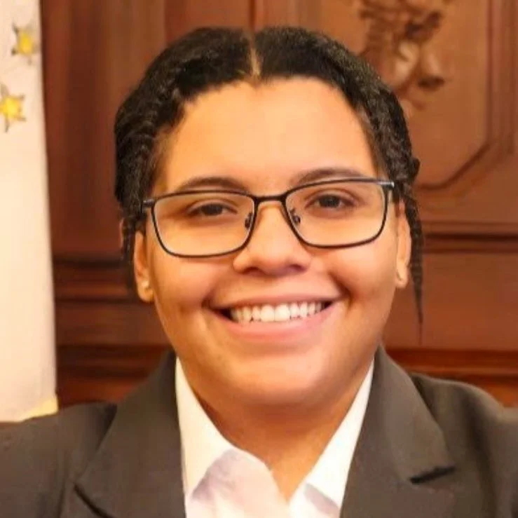 Portrait of a woman with short dark hair wearing glasses and a gray suit with white shirt, smiling at the camera in what appears to be an indoor setting with wooden paneling in the background.