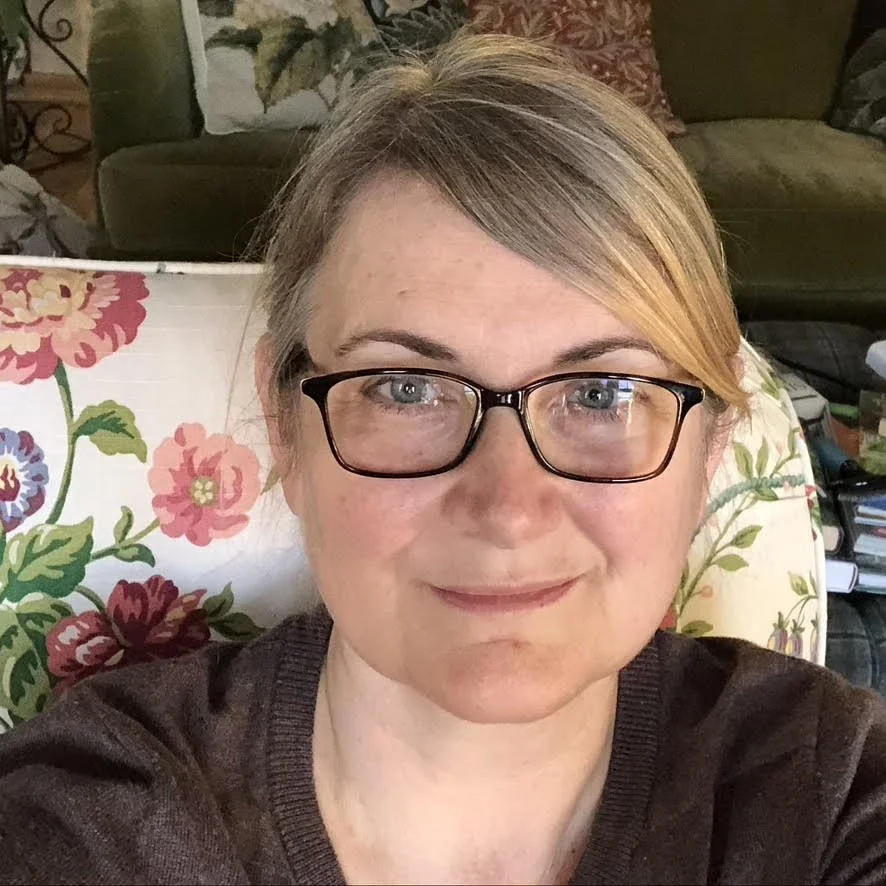 Casual portrait of a woman with blonde hair wearing glasses and a gray sweater, sitting in a floral-patterned chair in what appears to be a home setting.