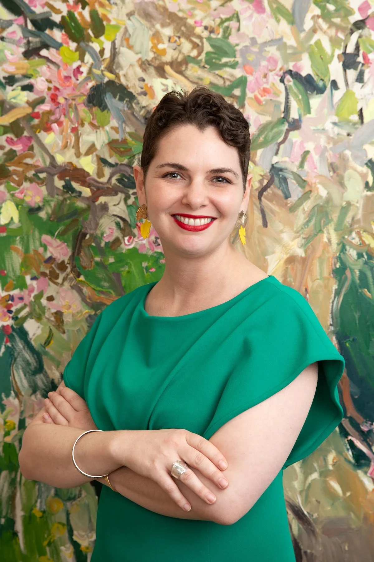 Professional headshot of a woman with short brown hair wearing a bright green dress and yellow earrings, smiling at the camera with arms crossed, standing in front of a colorful abstract floral painting
