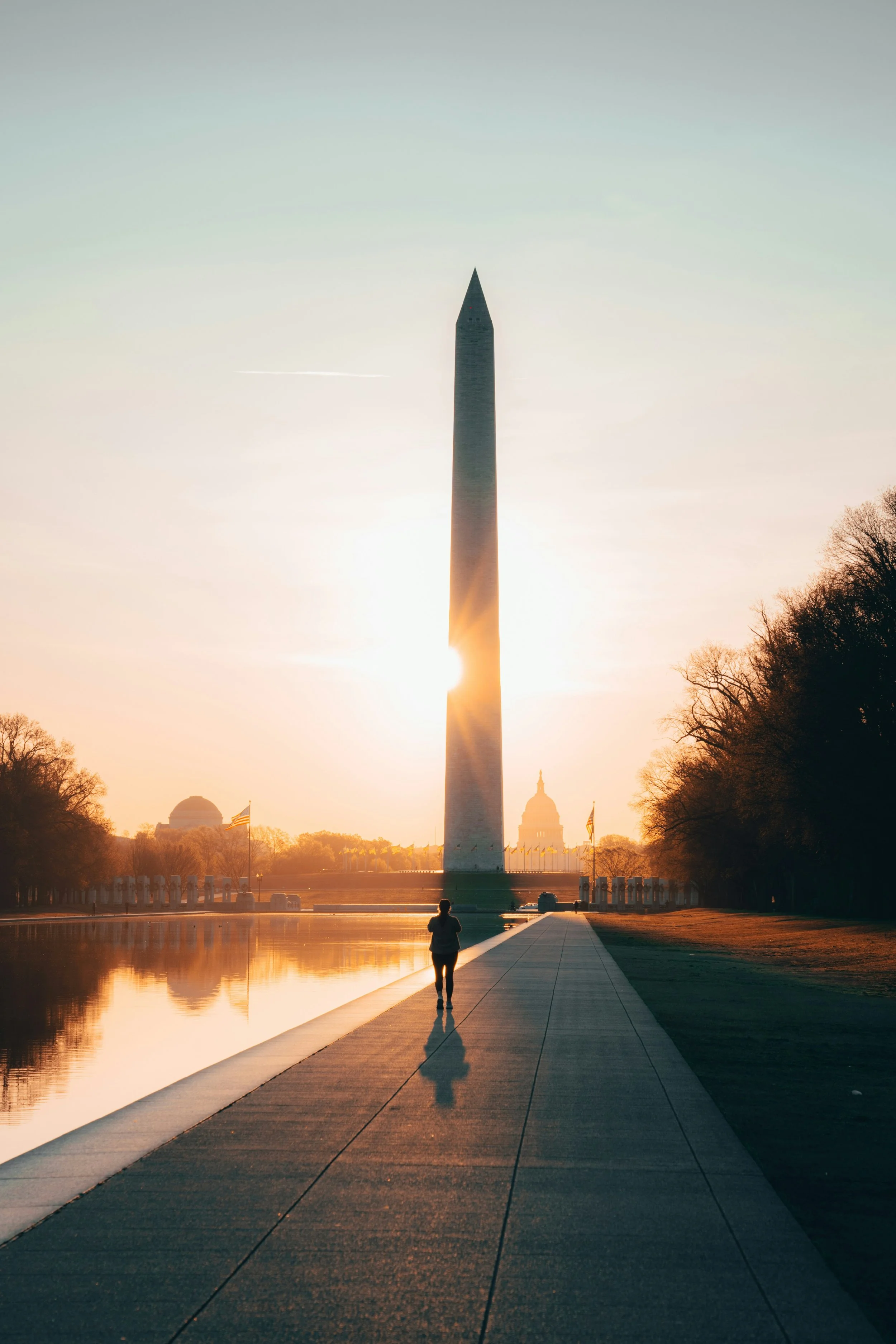 The Washington Monument at sunrise or sunset, with a person jogging along the reflecting pool walkway, and the U.S. Capitol building in the background.