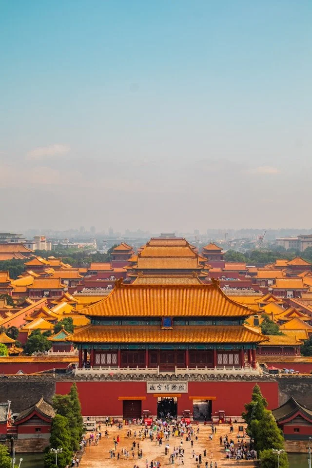 A large traditional Chinese palace with orange-tiled roofs and red walls, filled with crowds of tourists, under a clear blue sky.