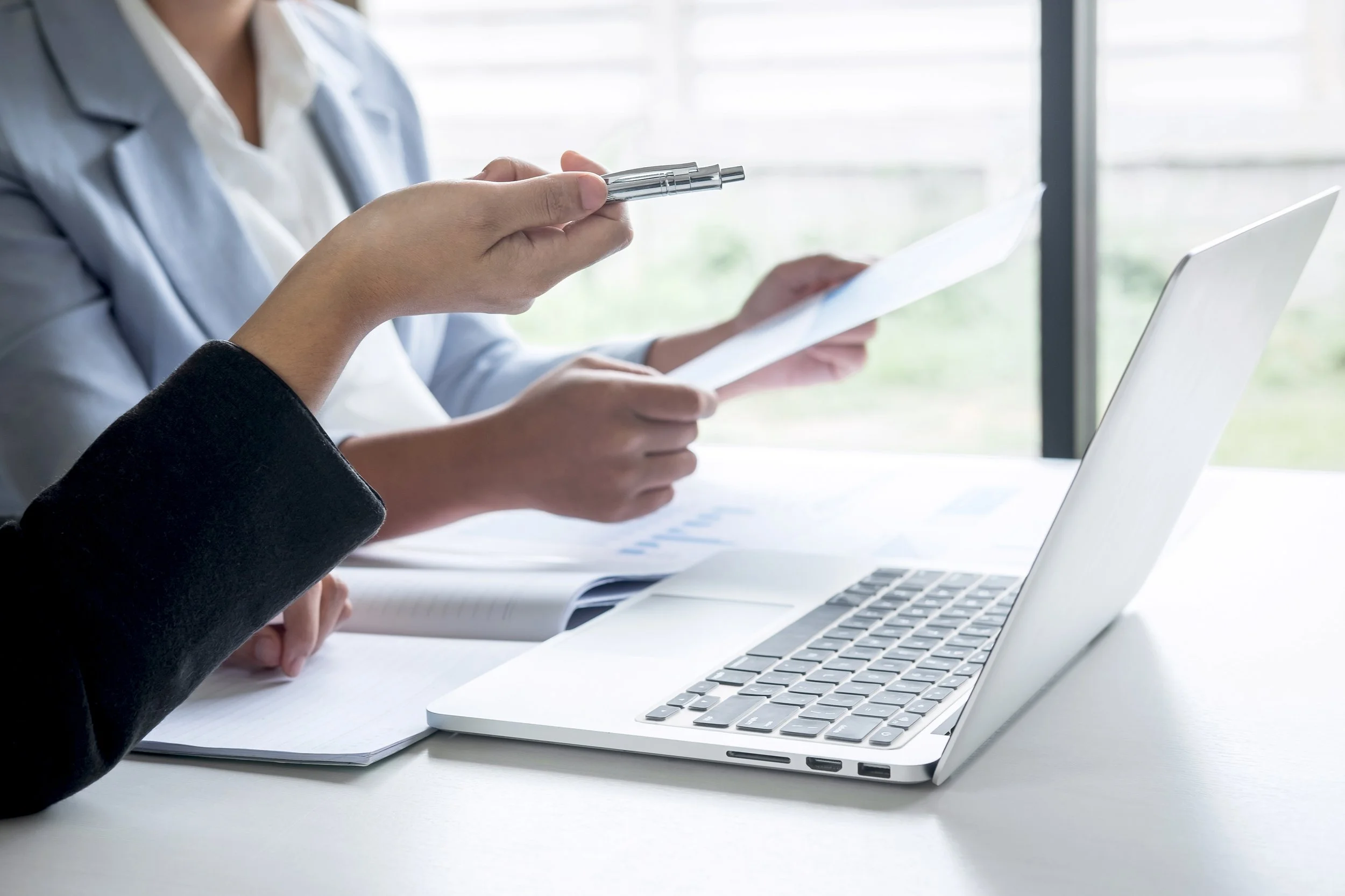 Two people in business attire working at a desk with a laptop, papers, and a pen, near a window.
