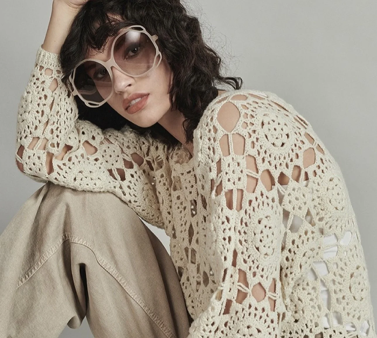 Woman with dark, curly hair wearing large, round sunglasses, a cream-colored crochet top, and beige pants, leaning her head on her arm.