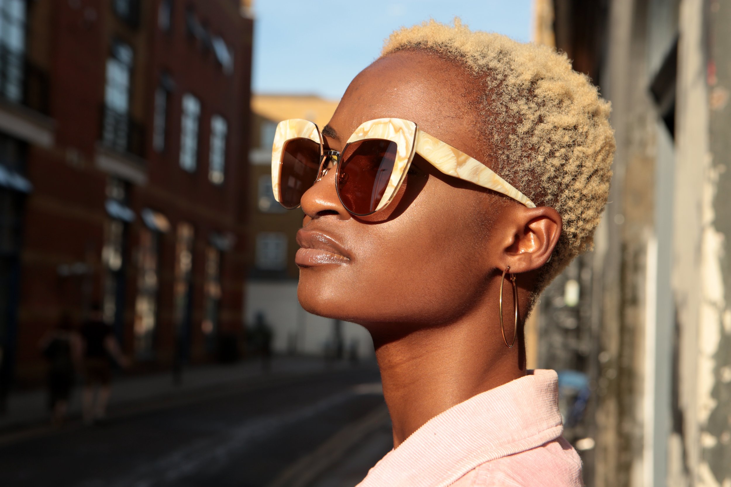 Close-up of a woman with short curly blonde hair, wearing large sunglasses with a unique beige frame, standing on a city street at sunset.