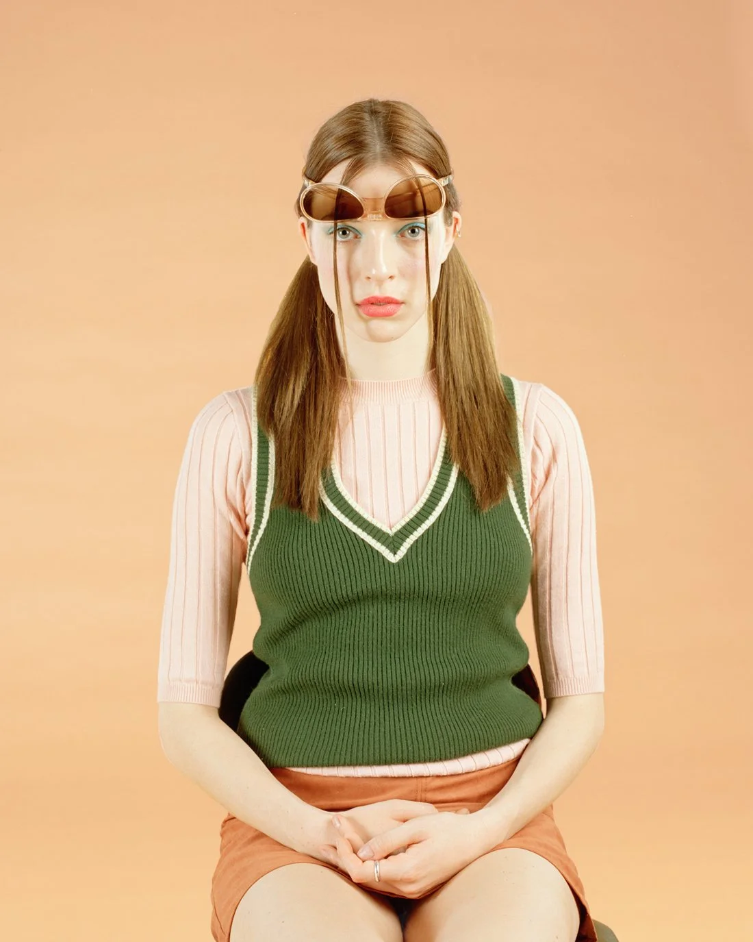 A young woman with long brown hair in pigtails, wearing sunglasses on her forehead, a pink and green layered sweater, and a peach-colored skirt, sitting against a beige background, with a surprised or confused expression.