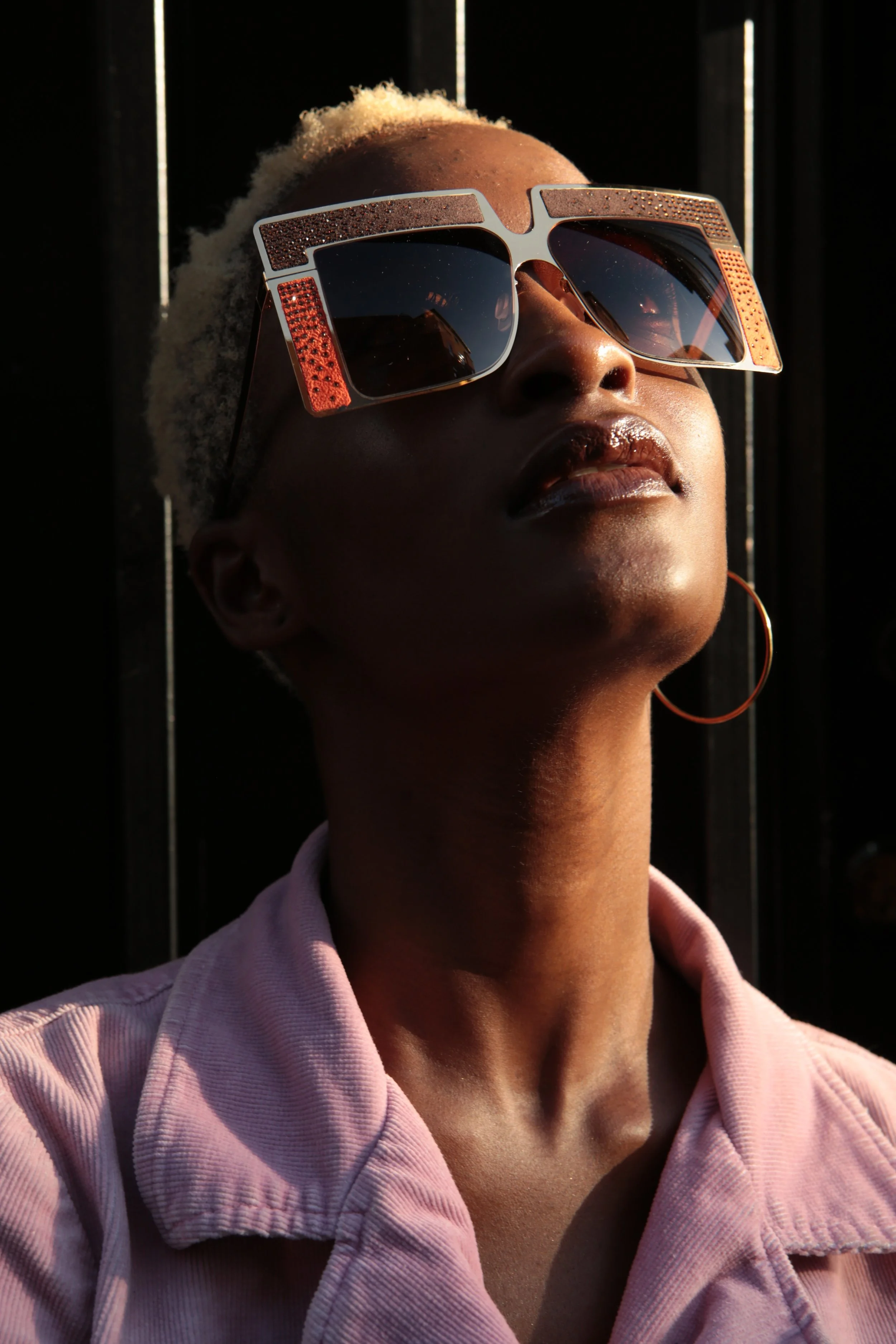 Portrait of a woman with short blonde hair wearing oversized sunglasses and hoop earrings, dressed in a pink shirt, facing slightly upward with sunlight highlighting her face.