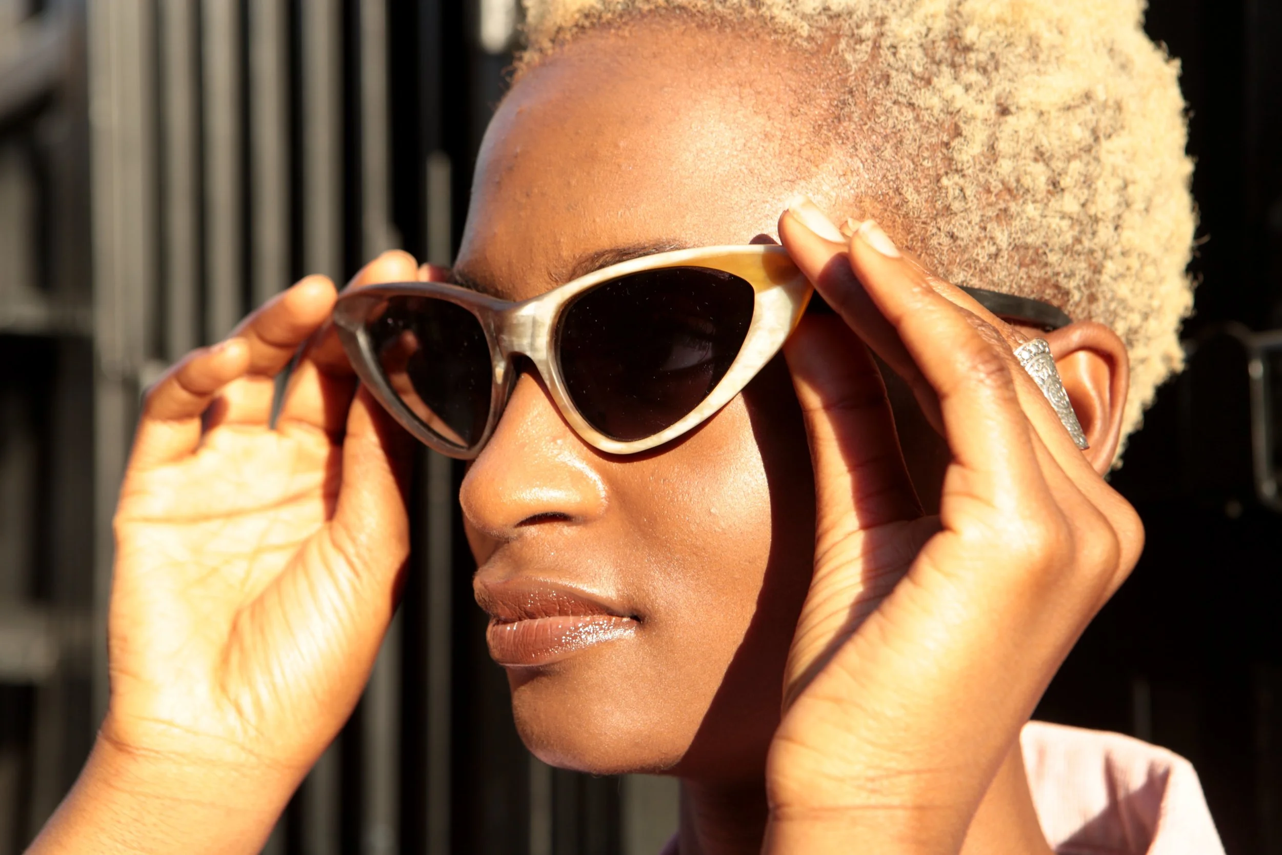 A woman with short, blonde curly hair wearing sunglasses and adjusting them with both hands.
