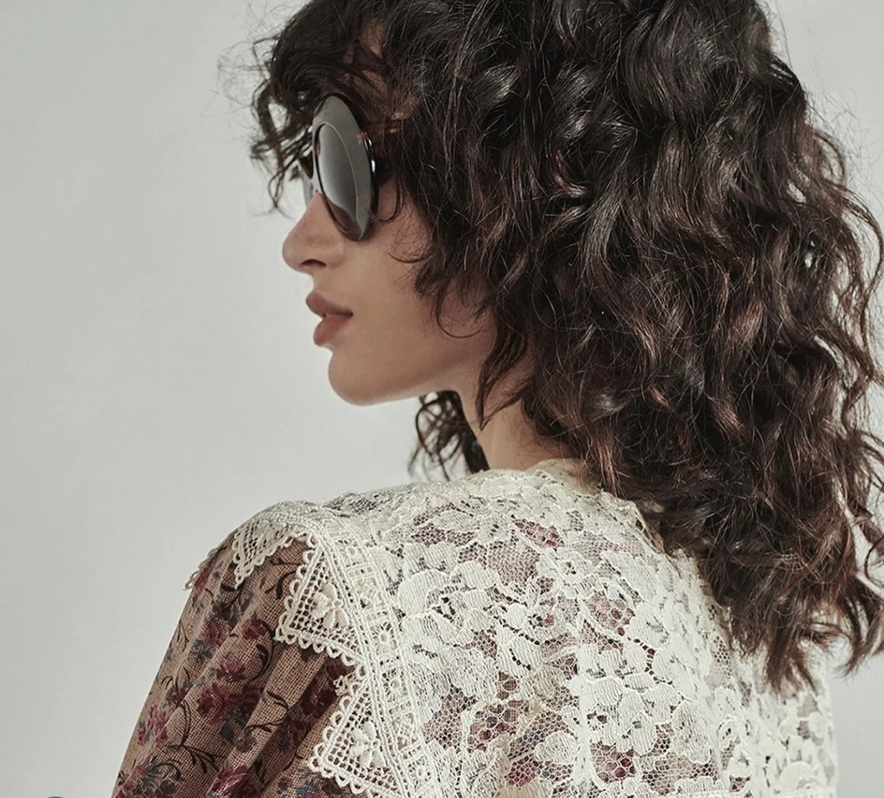 A woman with curly brown hair wearing large black sunglasses and a white lace top, looking to her left.