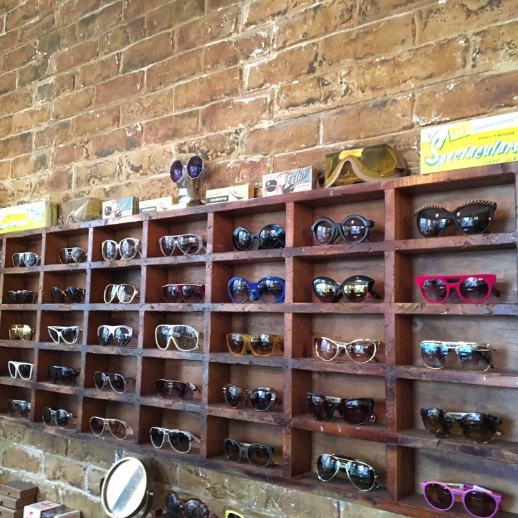 Display of various sunglasses on a wooden wall-mounted rack with a brick wall background.