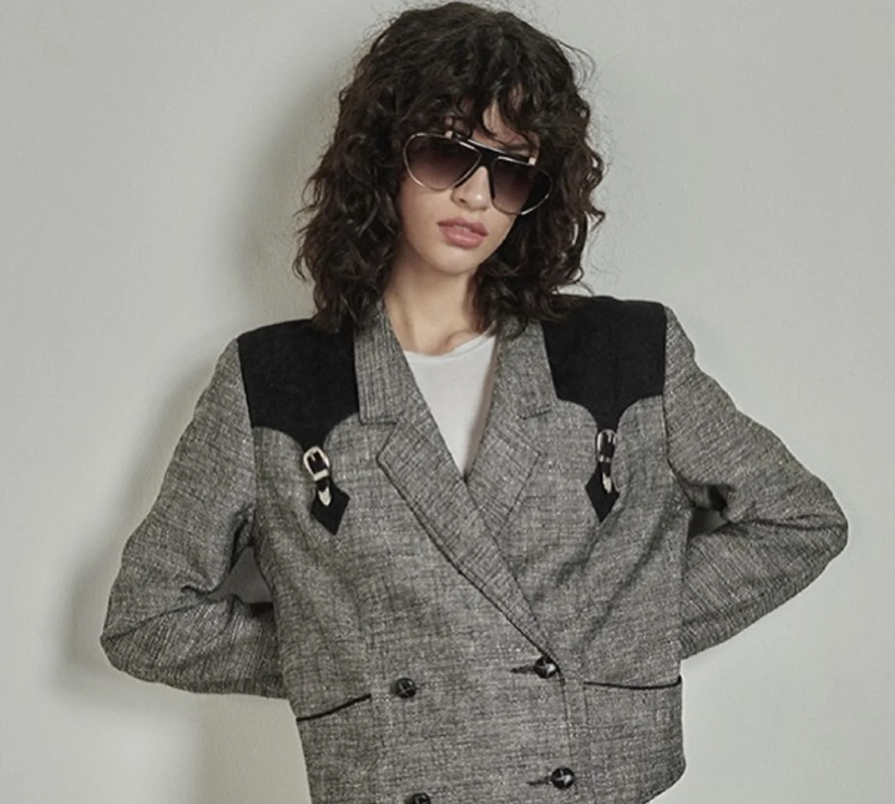 A woman with curly dark hair wearing large sunglasses, a white t-shirt, and a gray checked blazer with black shoulder patches and buckle details, posing against a plain light-colored wall.