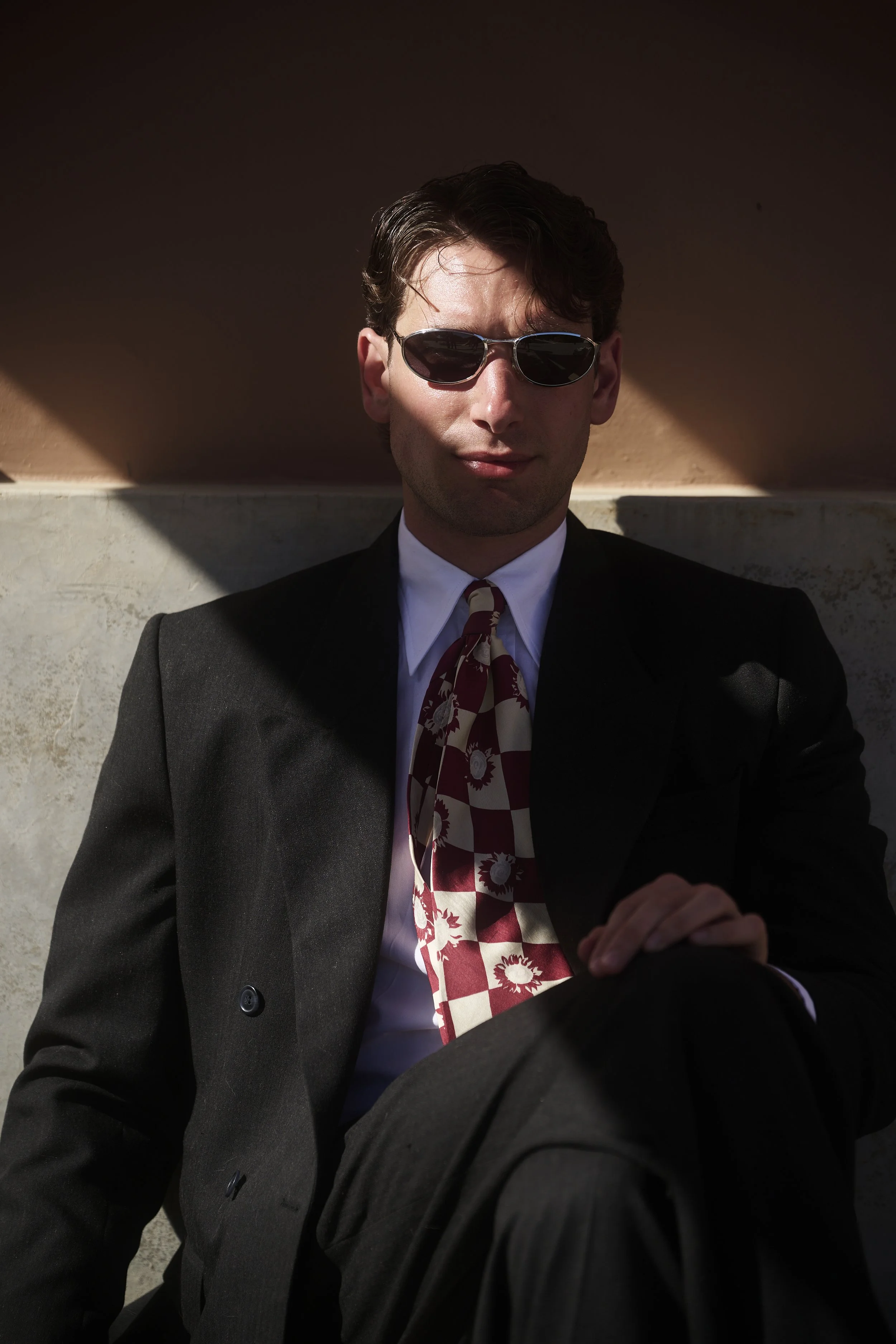 A man in a suit and tie, wearing sunglasses, sitting against a wall in sunlight.
