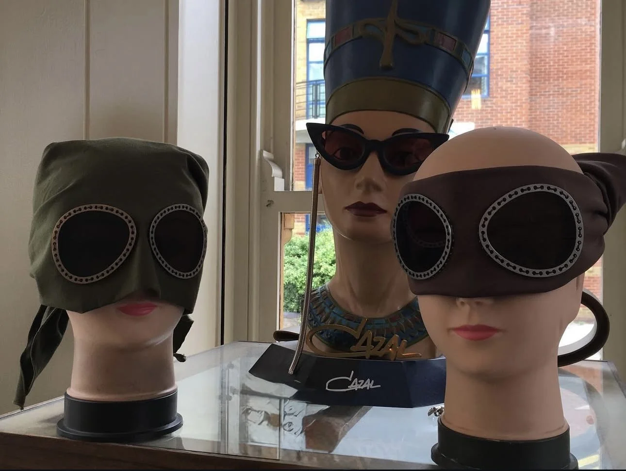 Three mannequin heads wearing different styles of sunglasses with two of them also wearing head coverings, displayed on a glass surface near a window.