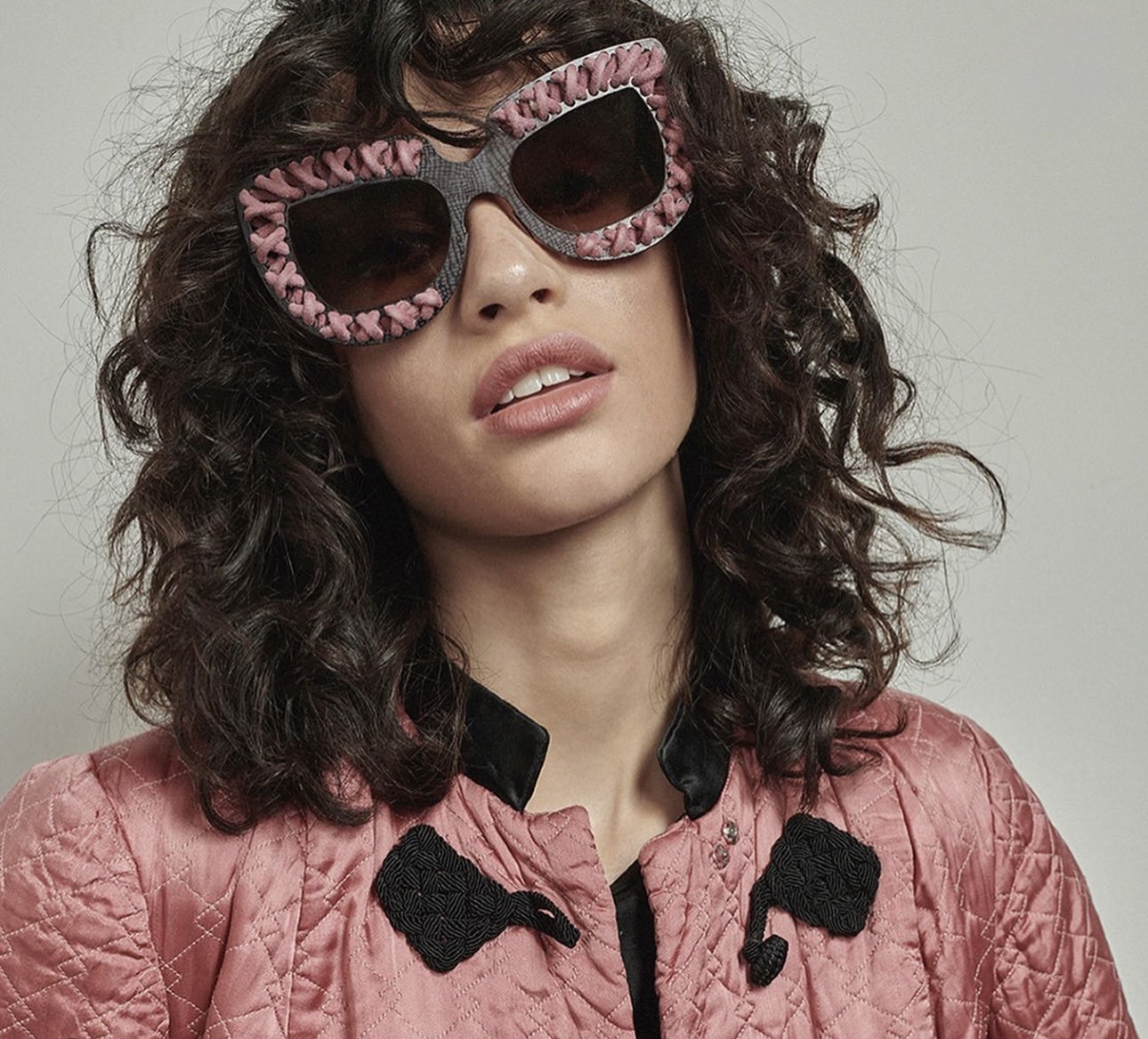 A woman with curly hair wearing heart-shaped sunglasses with pink and gray frames and dark lenses, and a pink quilted jacket with black bow embellishments.