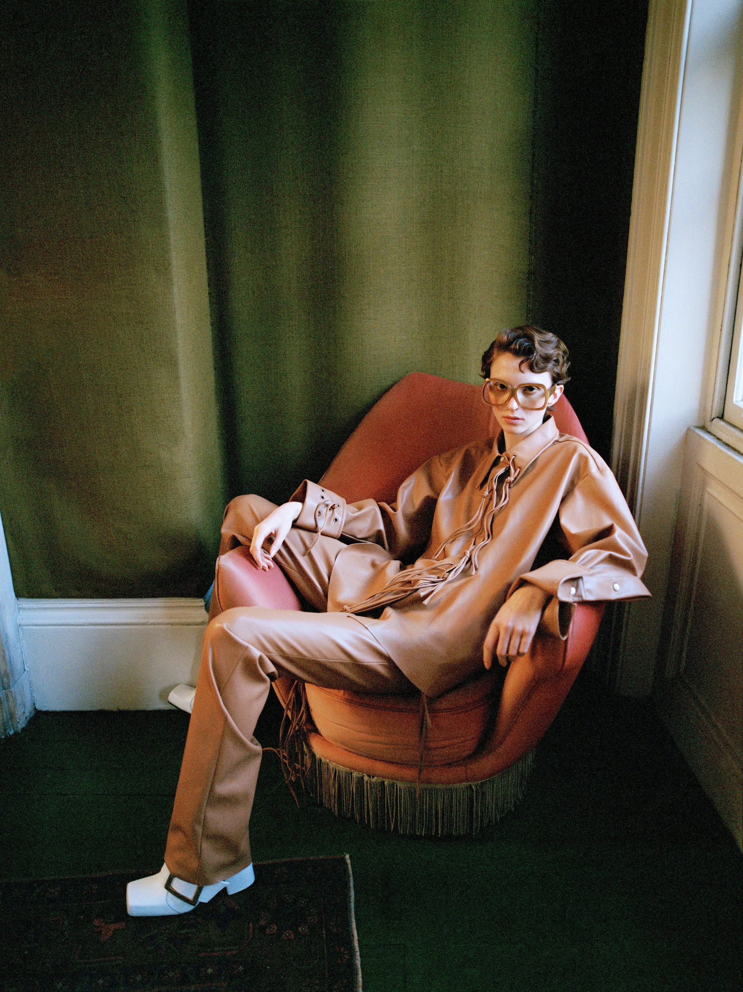 A young woman with short curly hair and glasses sits on a pink, round, vintage-style chair with fringed base, in a room with dark green walls and a large window nearby.