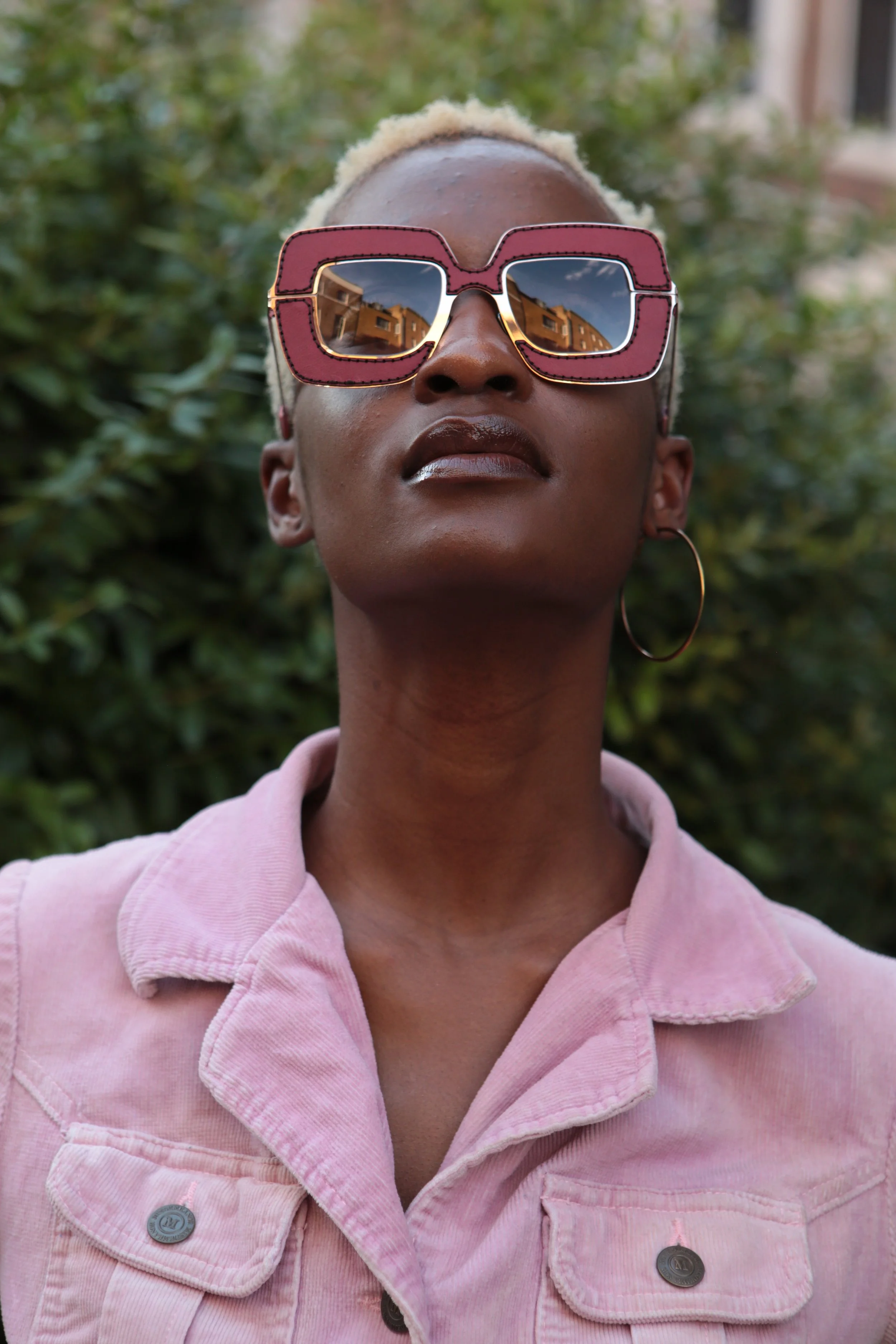 A woman with short blonde hair wearing large pink sunglasses with reflective lenses, a pink denim jacket, and hoop earrings, standing outdoors with green bushes in the background.